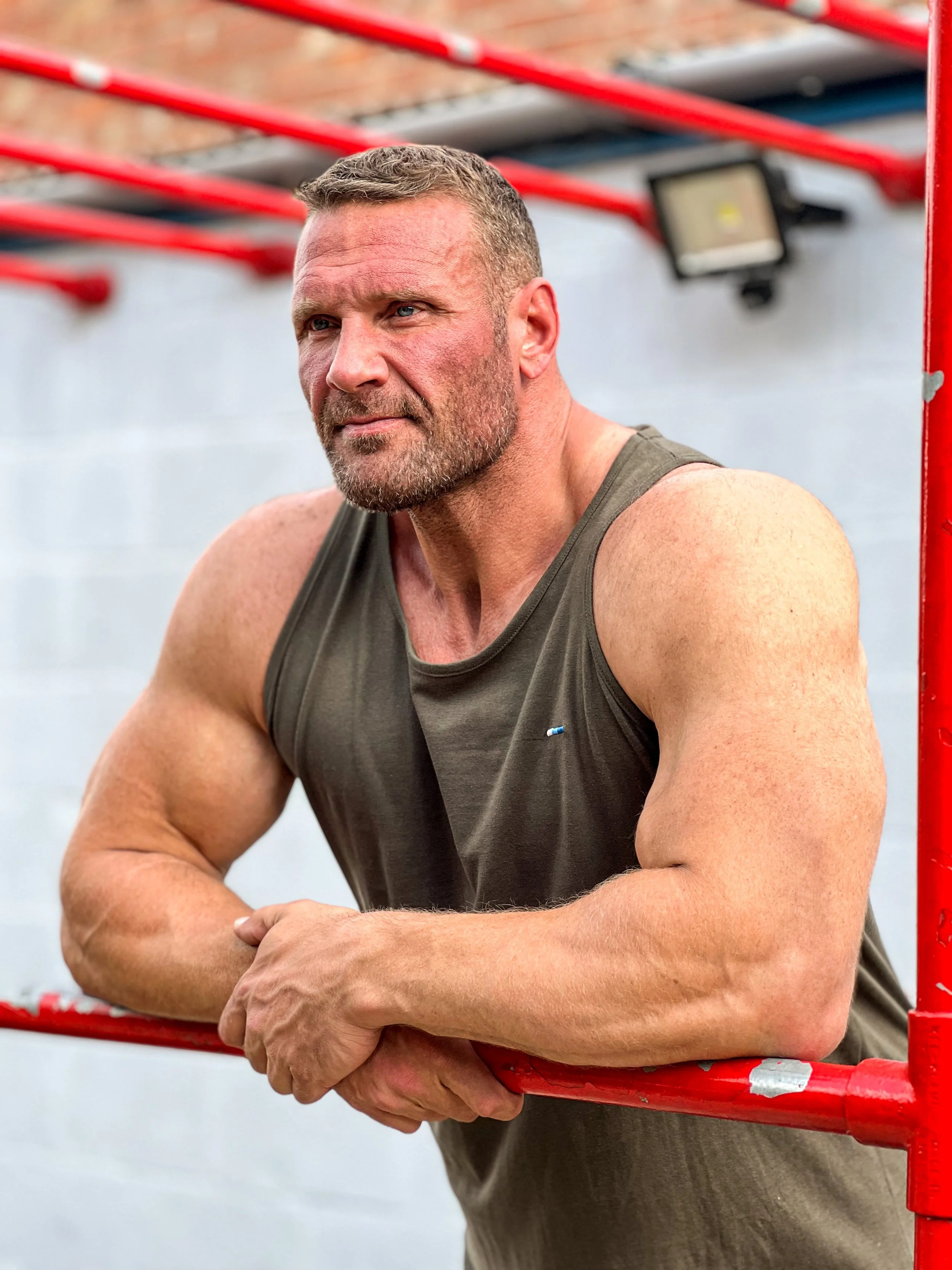 A muscular man with short hair and beard leaning on red gym equipment, wearing a sleeveless gray shirt, outdoors.