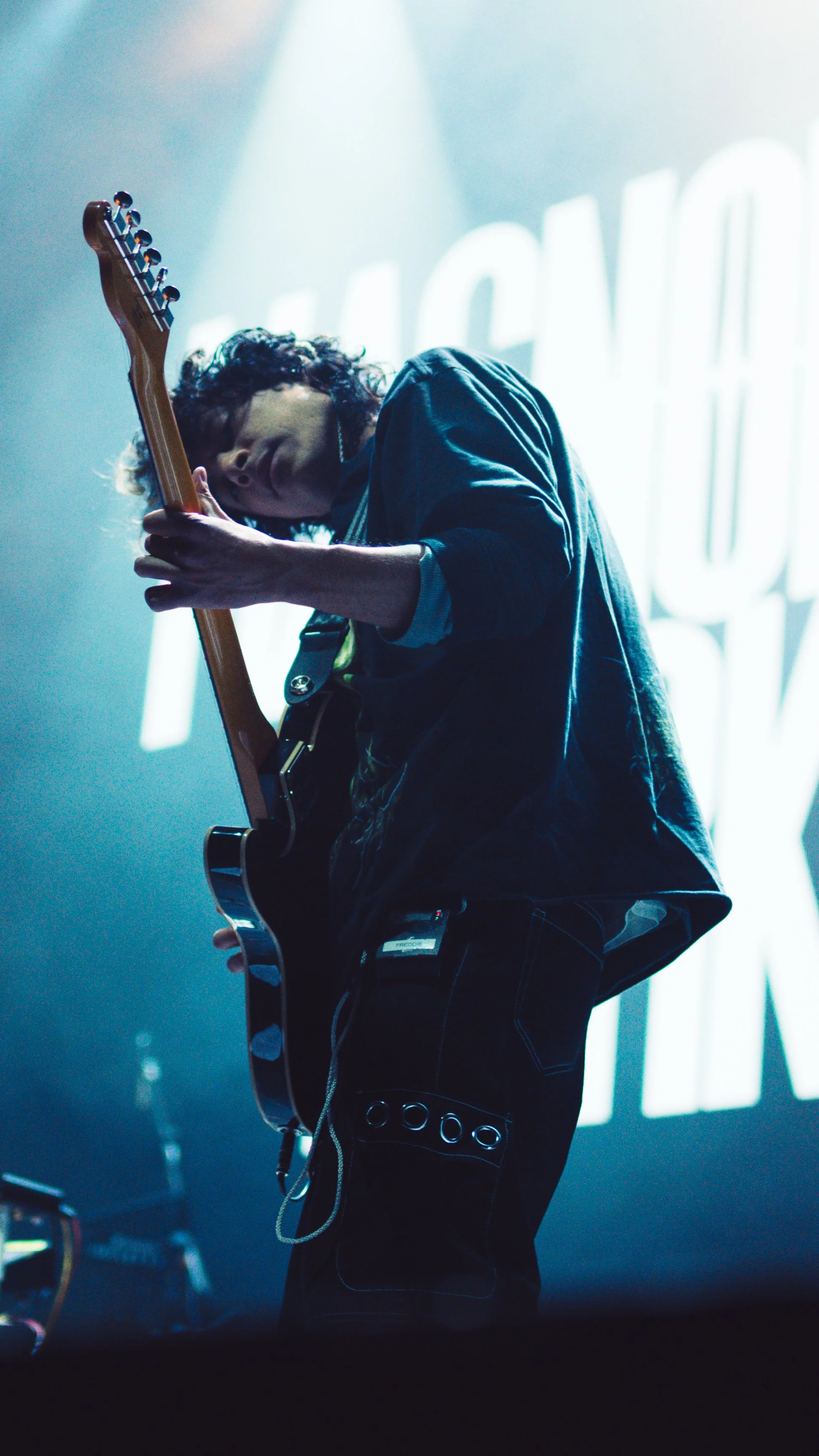 Musician playing electric guitar on stage with bright background lighting and blurred text behind him.