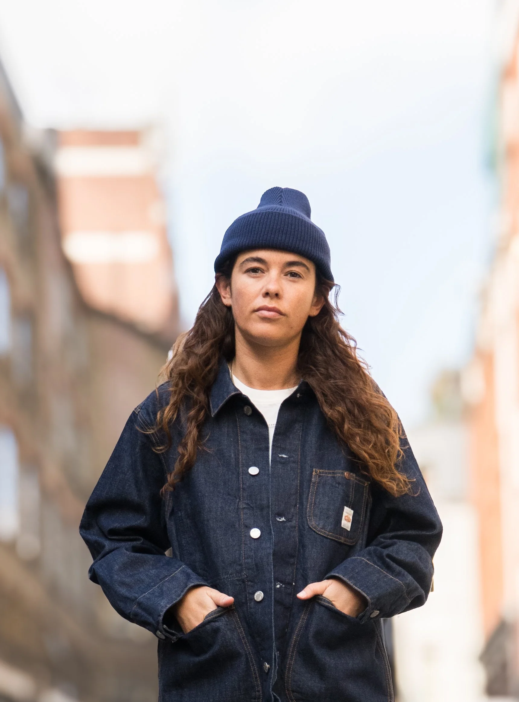 A woman with long, wavy brown hair wearing a dark denim jacket and navy knit beanie standing outdoors in a blurred city background.