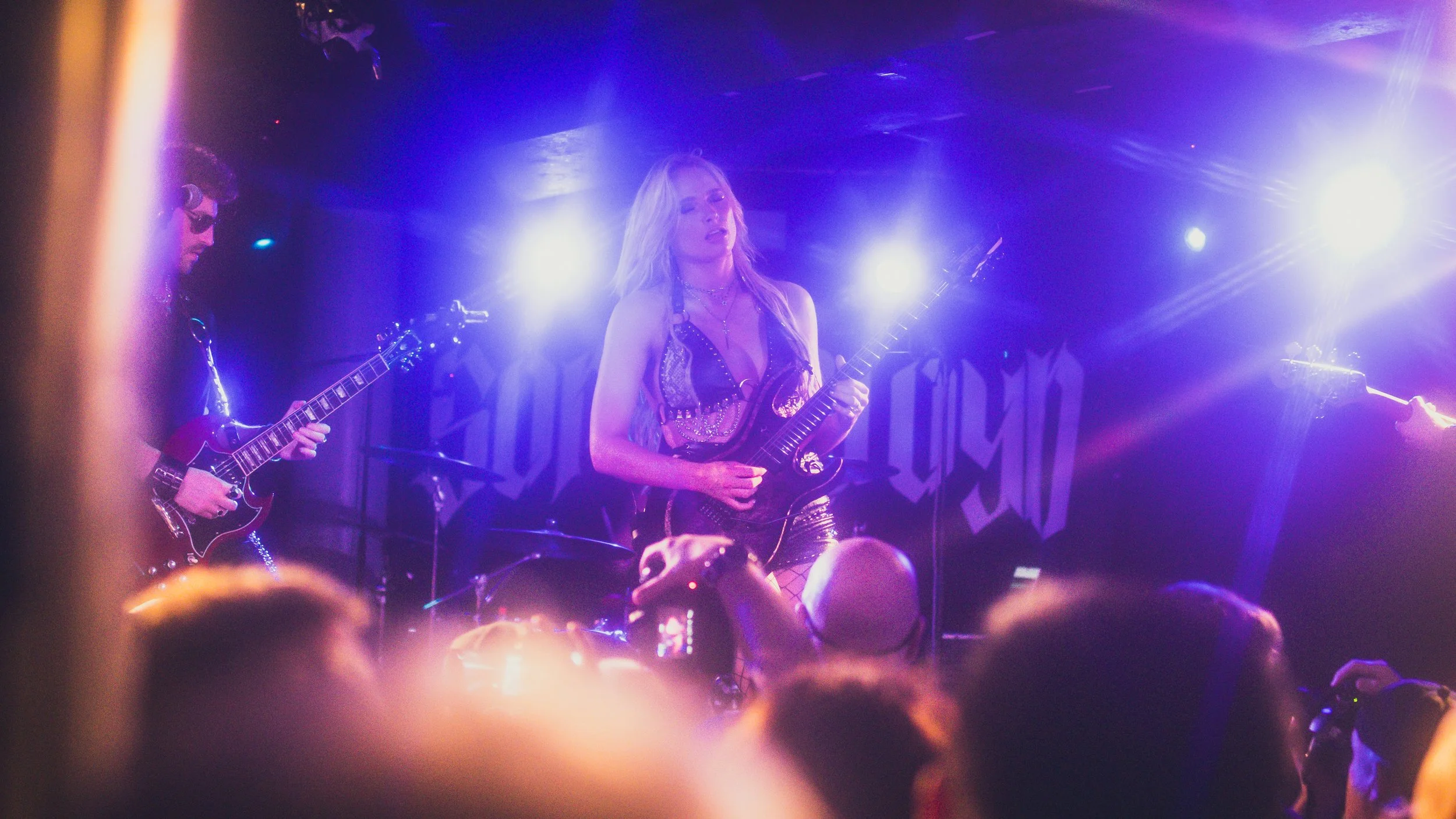 A woman playing an electric guitar on stage with purple and blue lighting, wearing a black and gold outfit, with an audience in front of her.