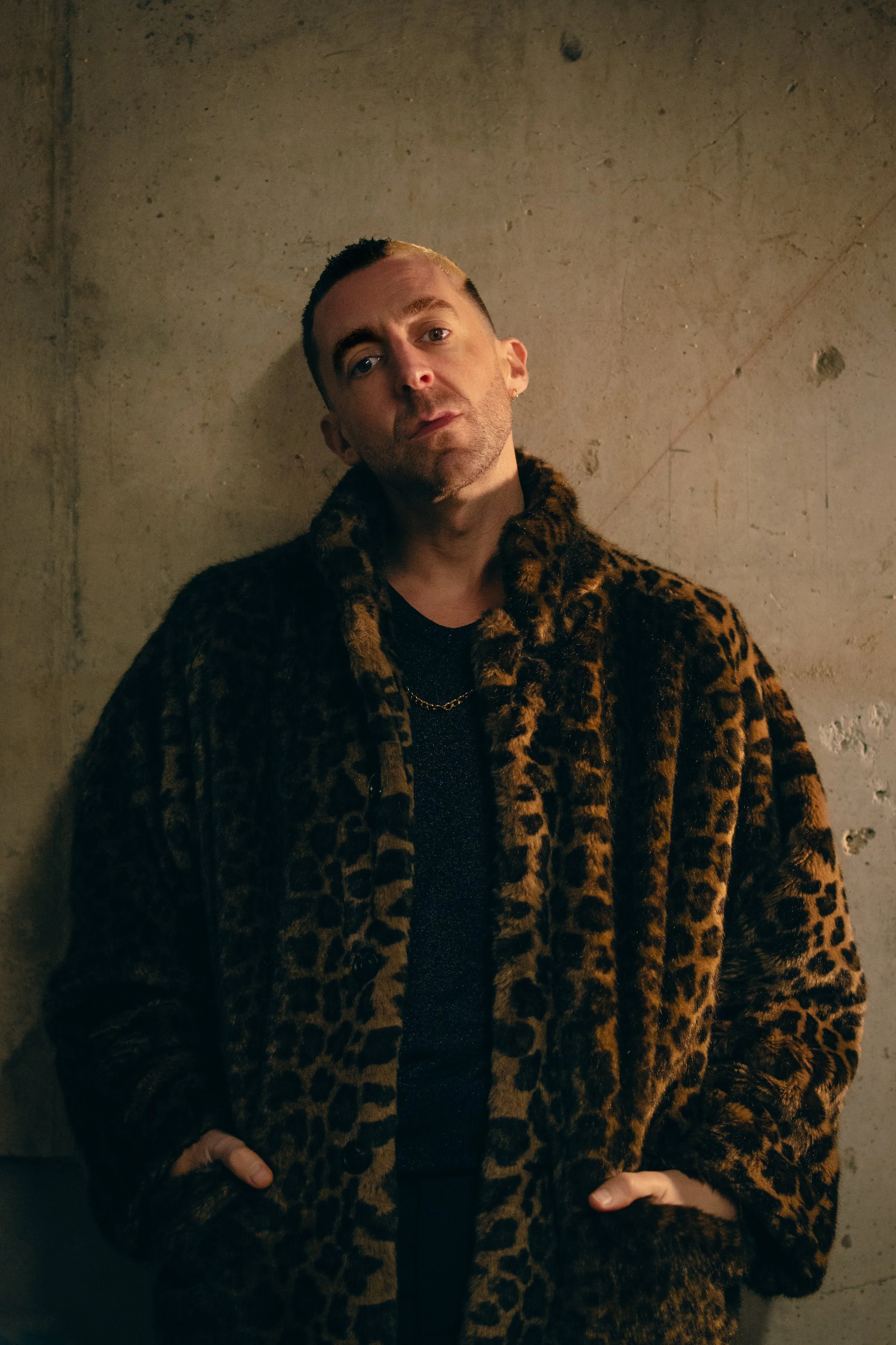 A man with dark hair and a shaved side, wearing a leopard print fur coat, standing against a textured beige wall.