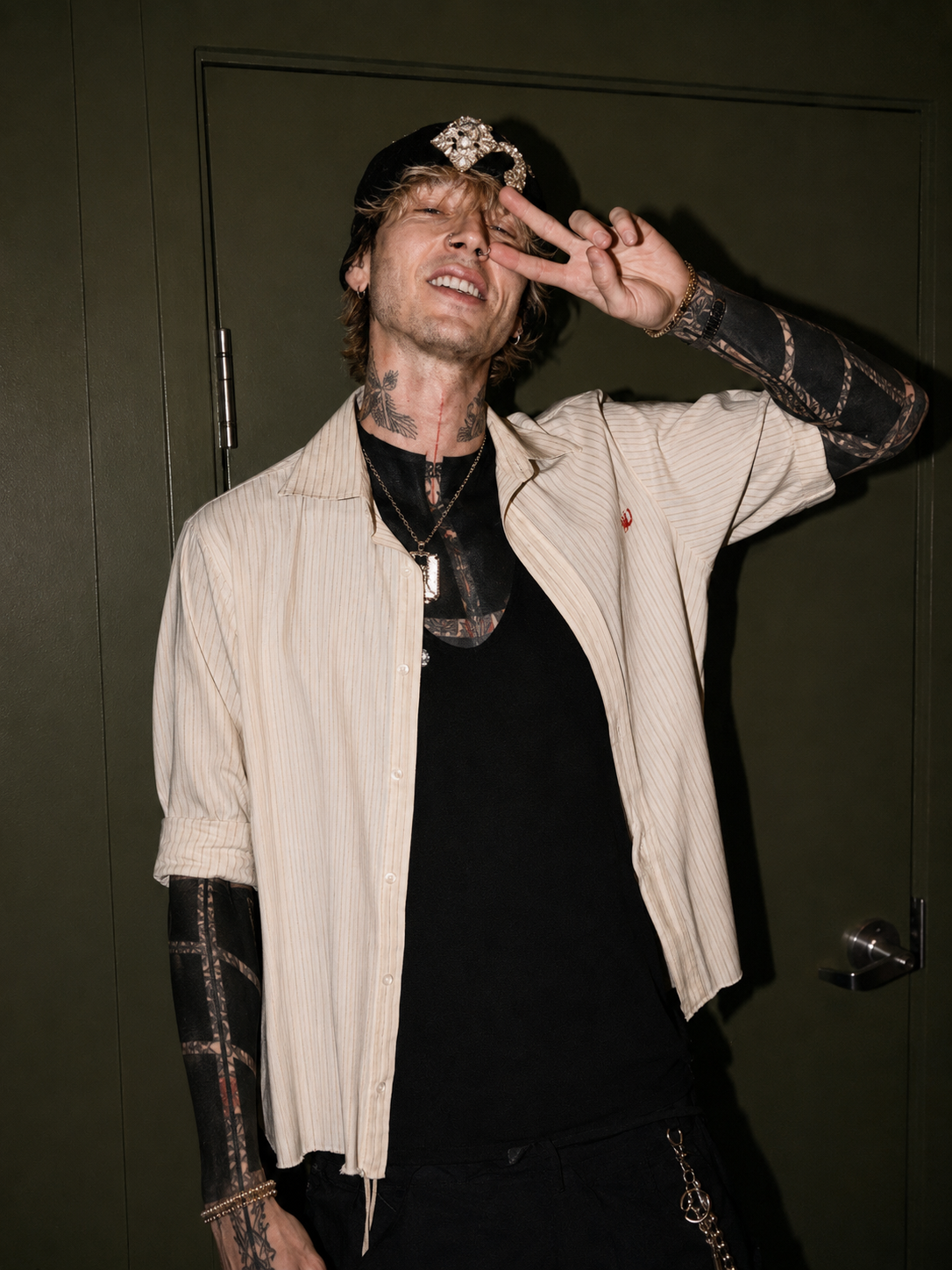 A man making a peace sign with his right hand near his eye, wearing a black cap with a jeweled brooch, striped beige shirt over a black t-shirt, and multiple tattoos visible on his neck and arms.