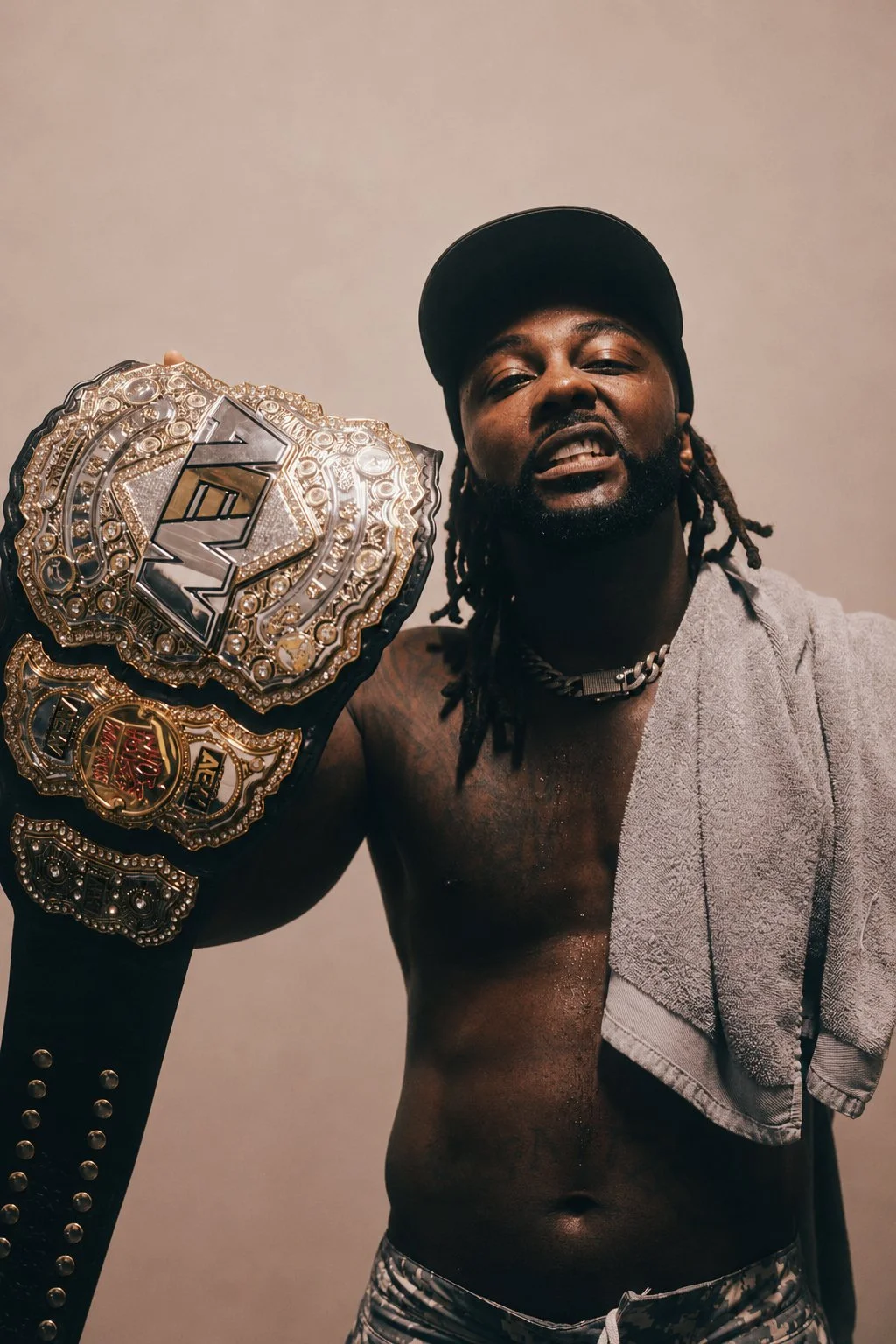 A shirtless man with dreadlocks and a black cap holding a large championship belt decorated with diamonds and gold. He has a towel draped over his shoulder and is wearing patterned shorts.