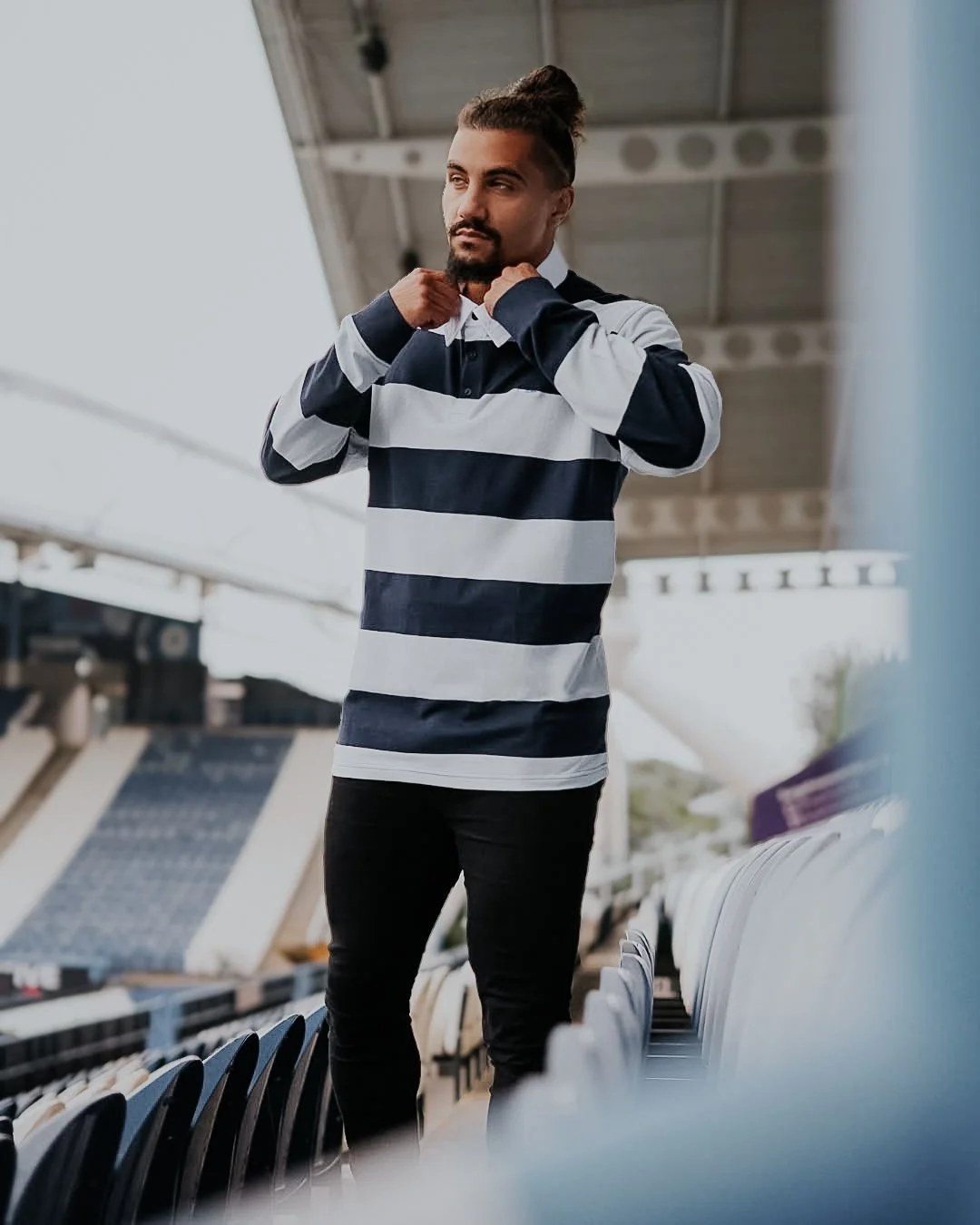 A young man adjusting his collar in a stadium with empty seats and a roof overhead, wearing a navy and white striped rugby shirt and black pants.
