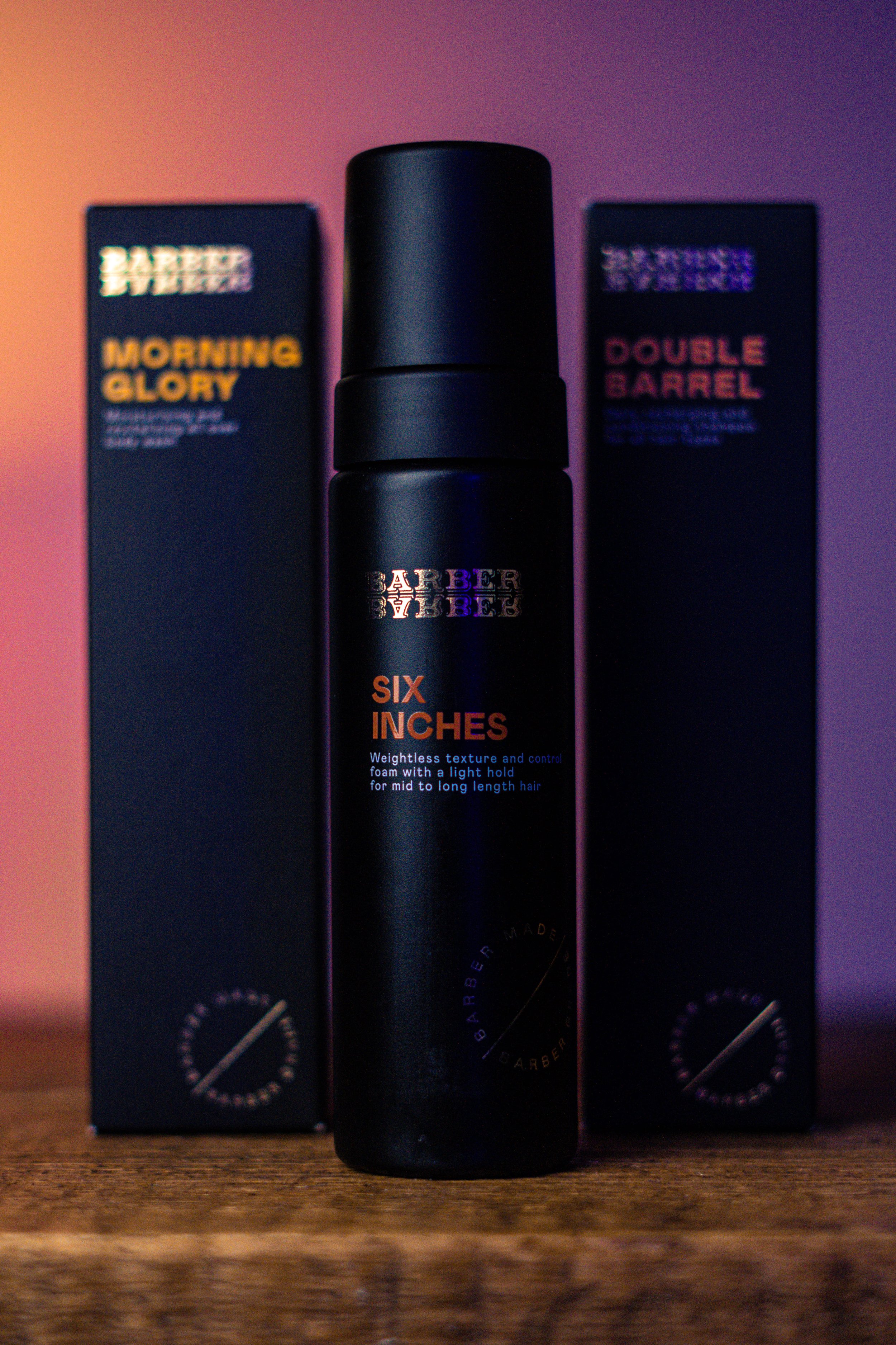 A black bottle of hair styling foam labeled 'Six Inches' in orange text, flanked by two boxes of hair products with purple and orange labels, on a wooden surface with colorful lighting.