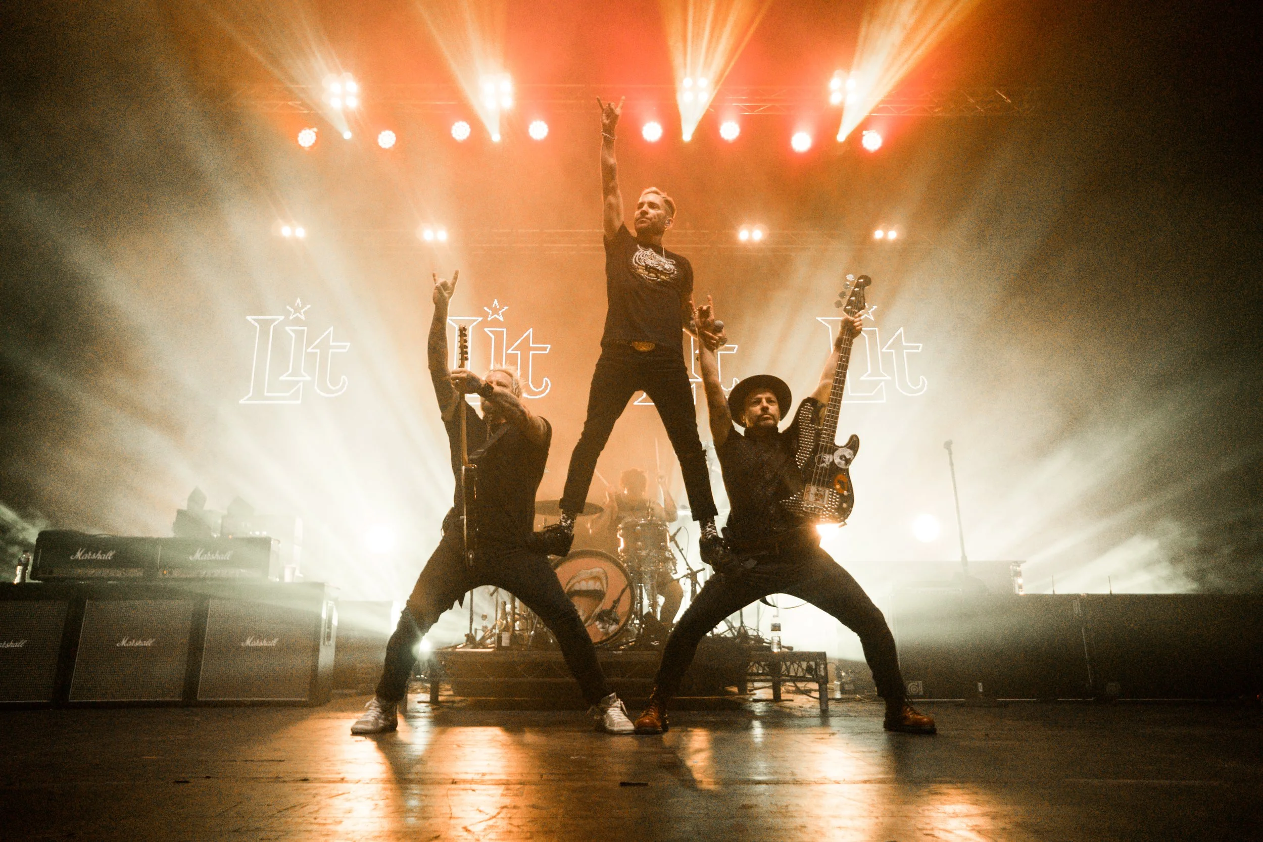 A band performing on stage with two guitarists and a vocalist, backlit by bright stage lights, in front of a illuminated logo reading 'Lit'.