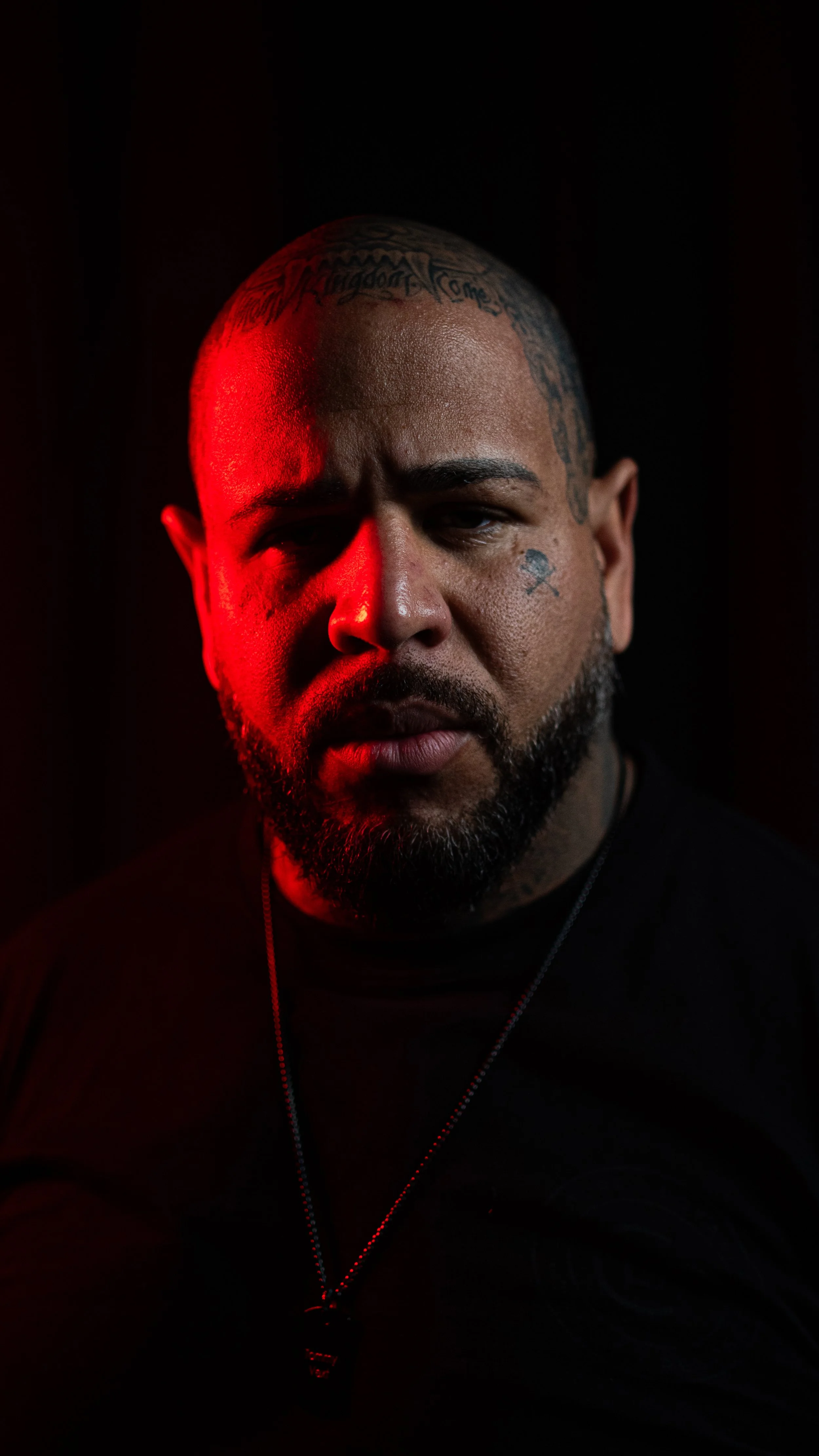 A man with a beard and tattoos on his face and head, illuminated by red and black lighting, wearing a black shirt and a necklace.