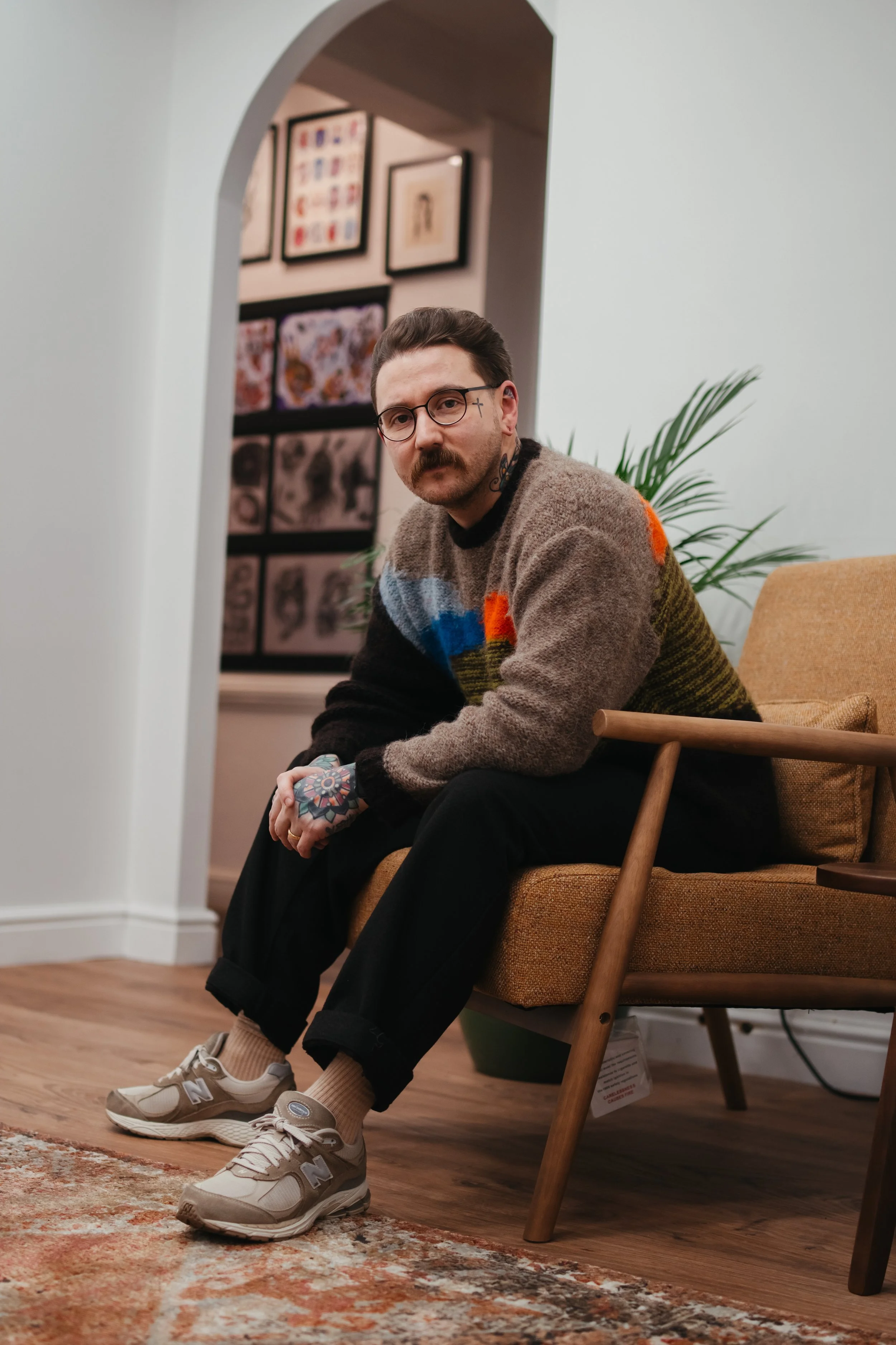 A man with glasses and a mustache, wearing a colorful knit sweater, black pants, and sneakers, sitting on a mustard-colored armchair inside a room with wooden flooring, indoor plants, and framed artwork on the wall behind him.