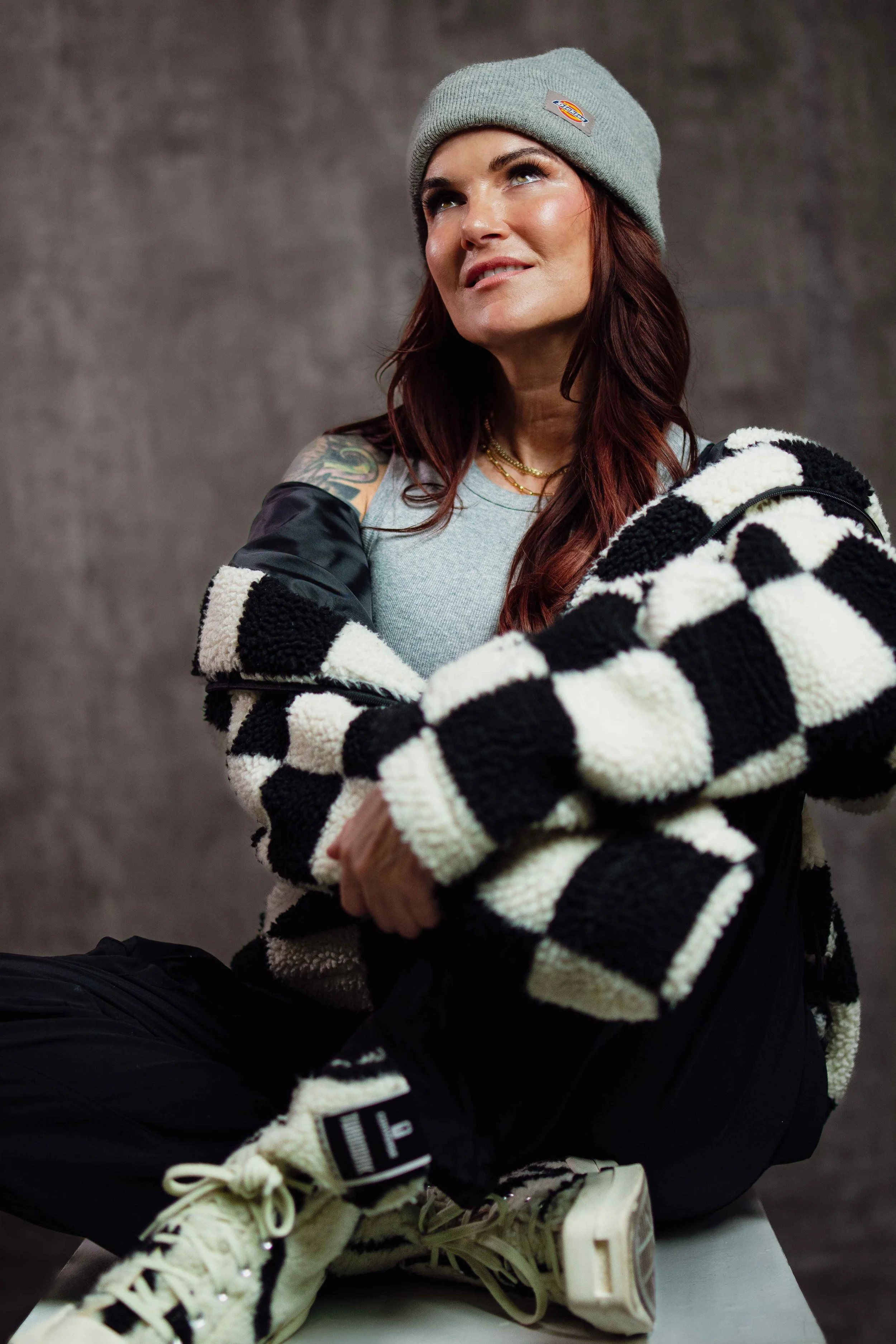 Woman with long hair wearing a gray beanie, colorful jewelry, a gray tank top, a black and white checkered jacket, black pants, and cream-colored sneakers, sitting against a gray background.