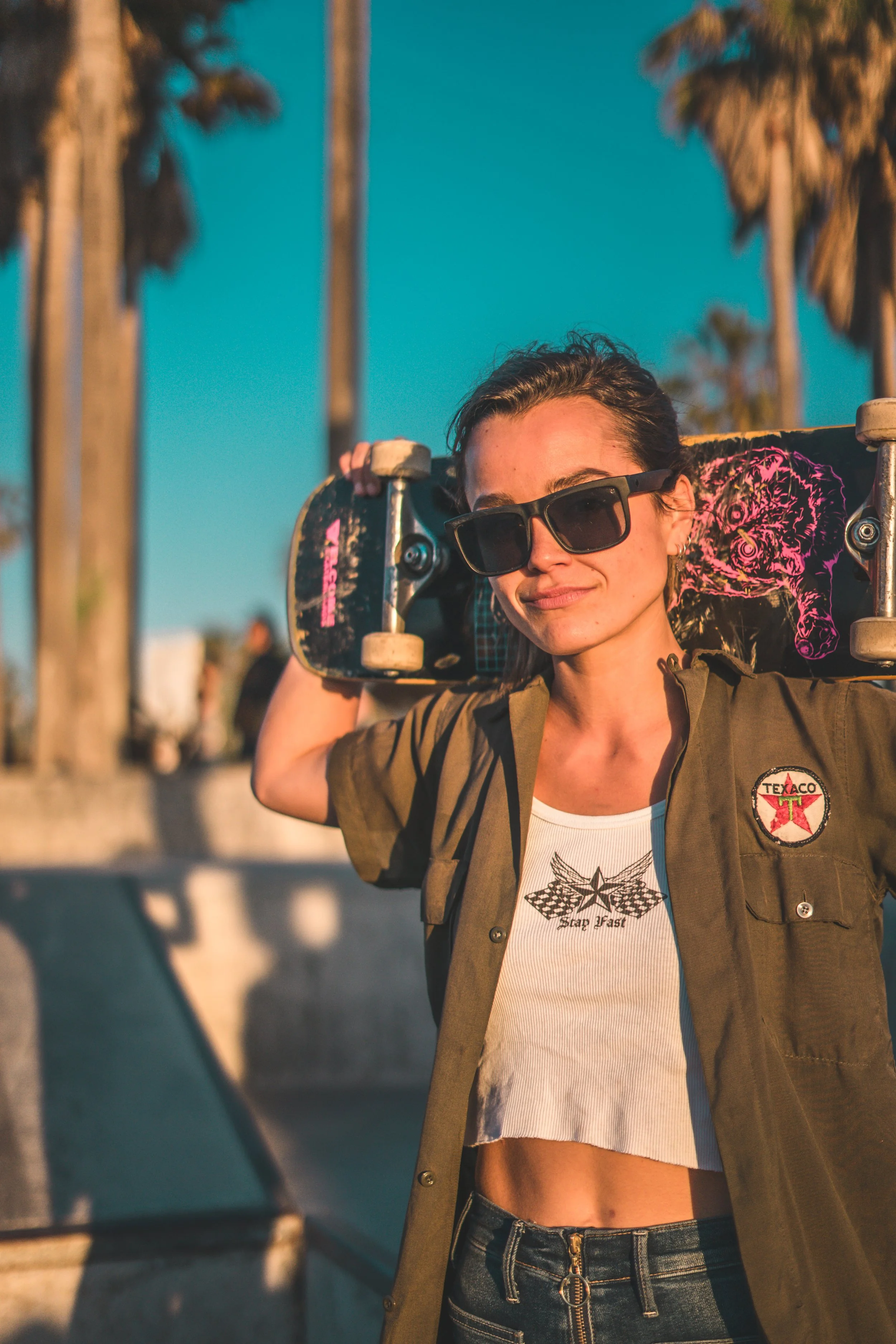 A woman with sunglasses and a casual outfit, holding a skateboard over her shoulder, standing outdoors in front of palm trees during sunset.