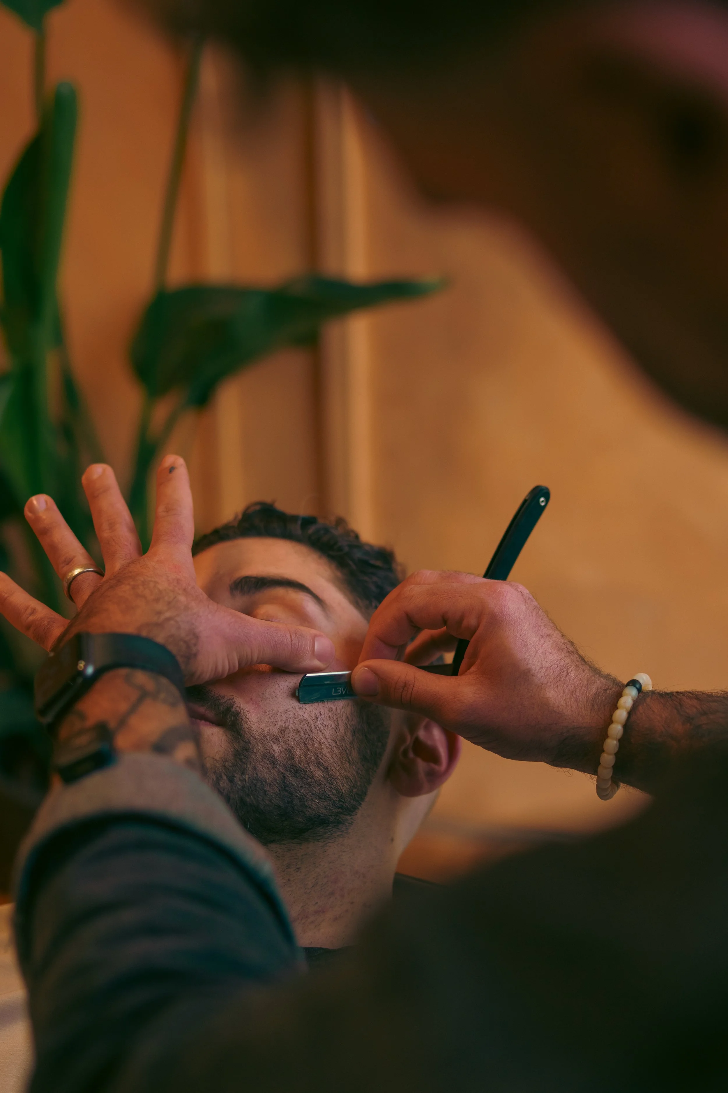 Close-up of a man getting a beard trim or shave by a barber using a straight razor in a barbershop with wooden paneling and a green plant in the background.