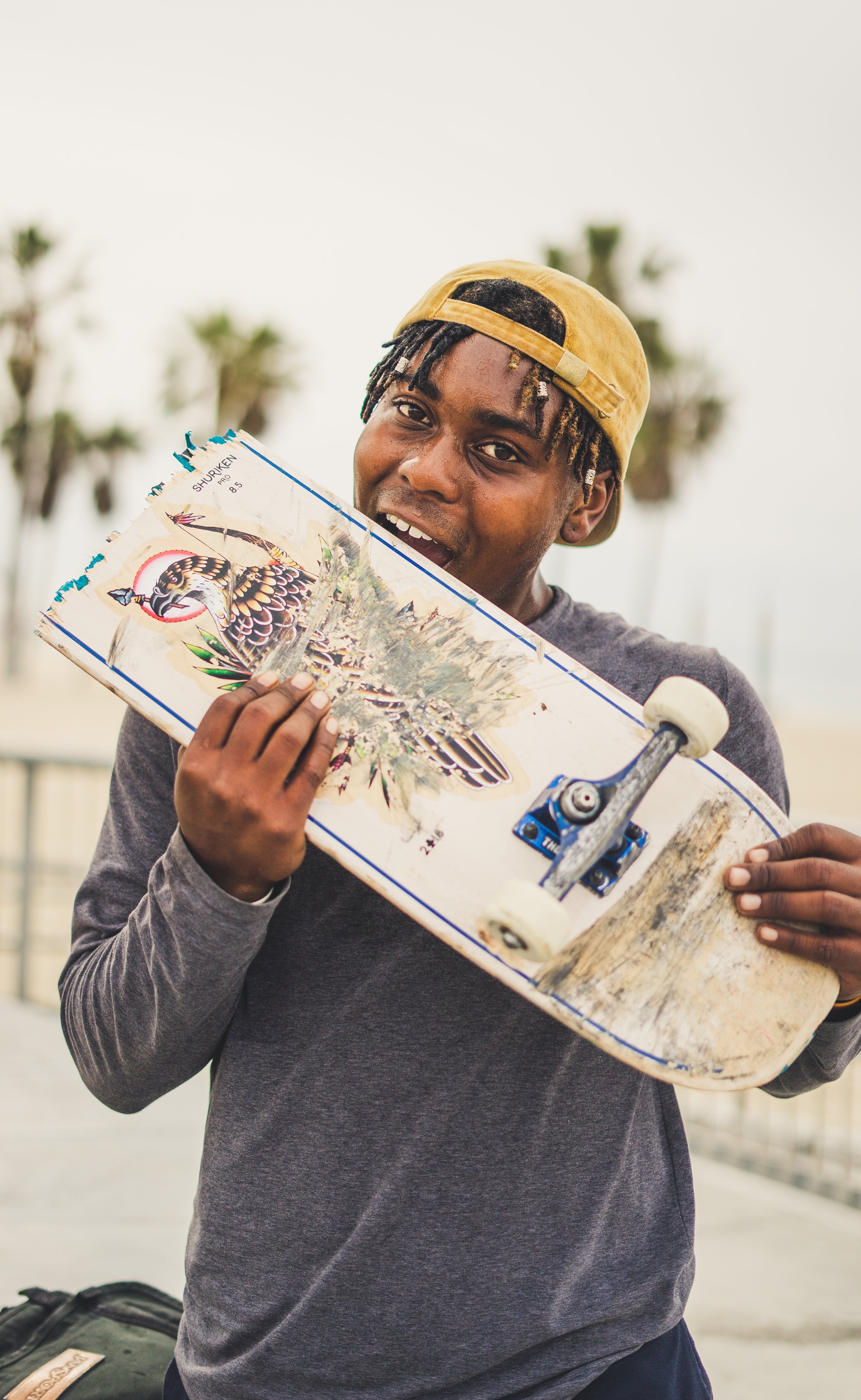 A young man holding a skateboard with a colorful bird illustration on it. He is wearing a yellow backward cap and a gray long sleeve shirt, standing outdoors with palm trees in the background.