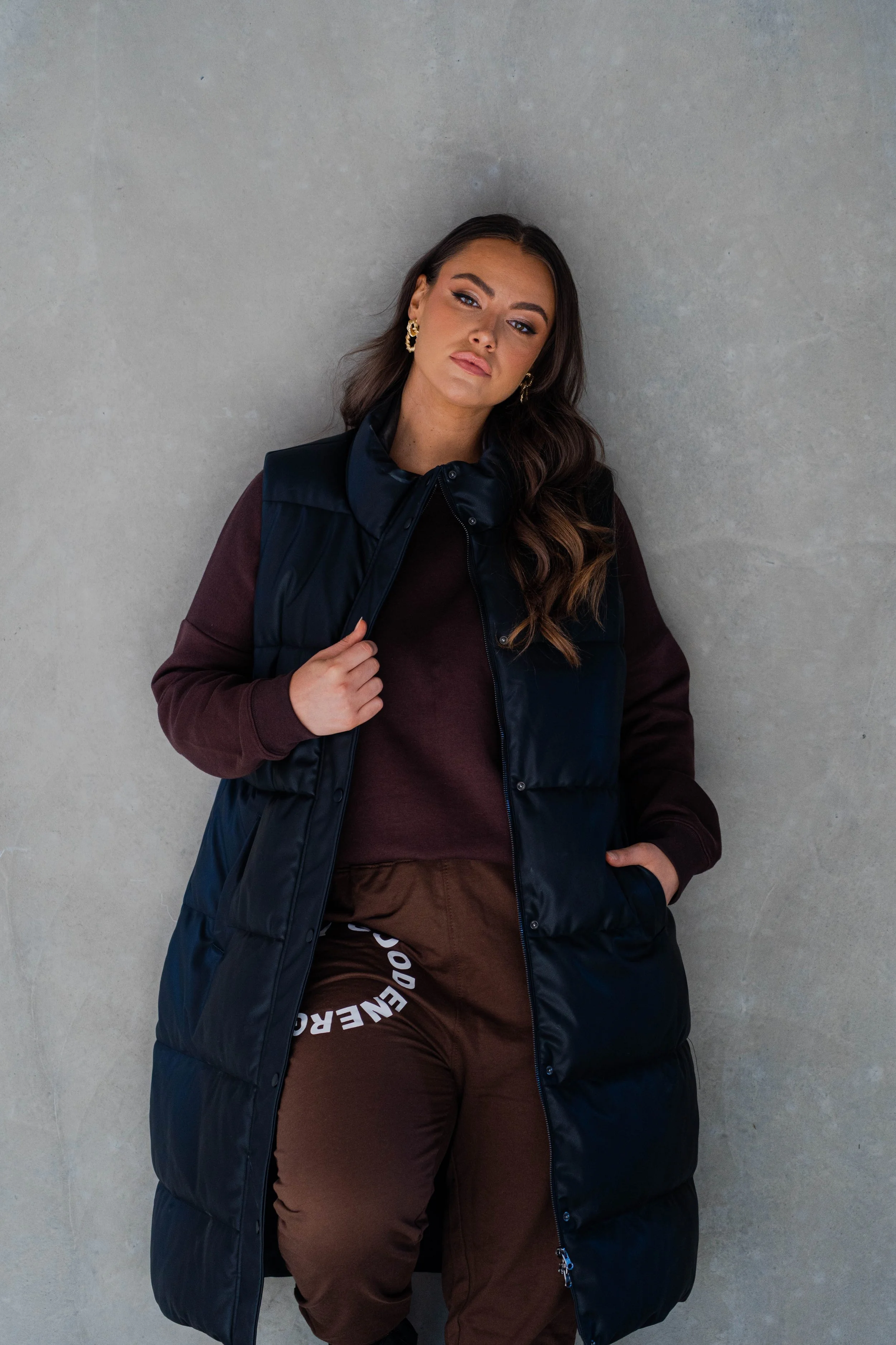 A woman with long wavy brown hair and gold earrings leaning against a plain gray wall, wearing a dark puffer vest over a maroon sweatshirt and brown pants.