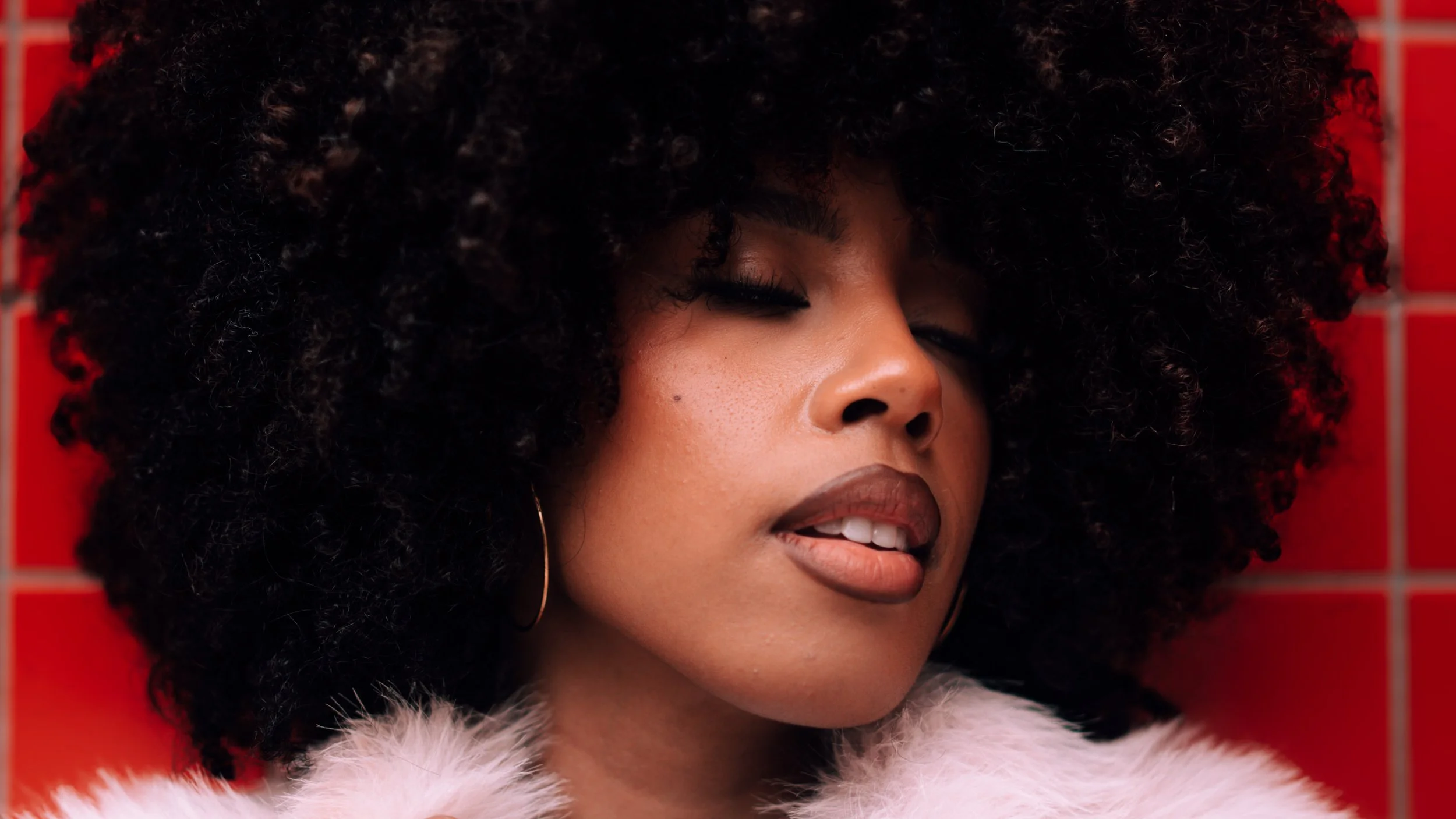 Close-up of a woman with curly hair, wearing makeup, a pink fluffy garment, and hoop earrings, against a red tiled background.