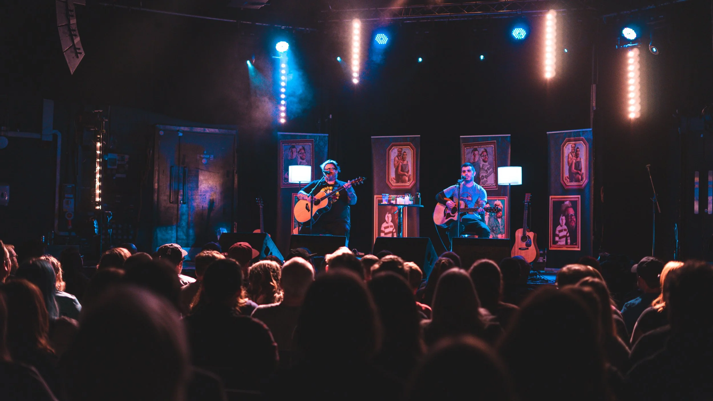 Two musicians are performing with guitars on a stage illuminated by blue and purple lighting, with audience watching in the foreground.