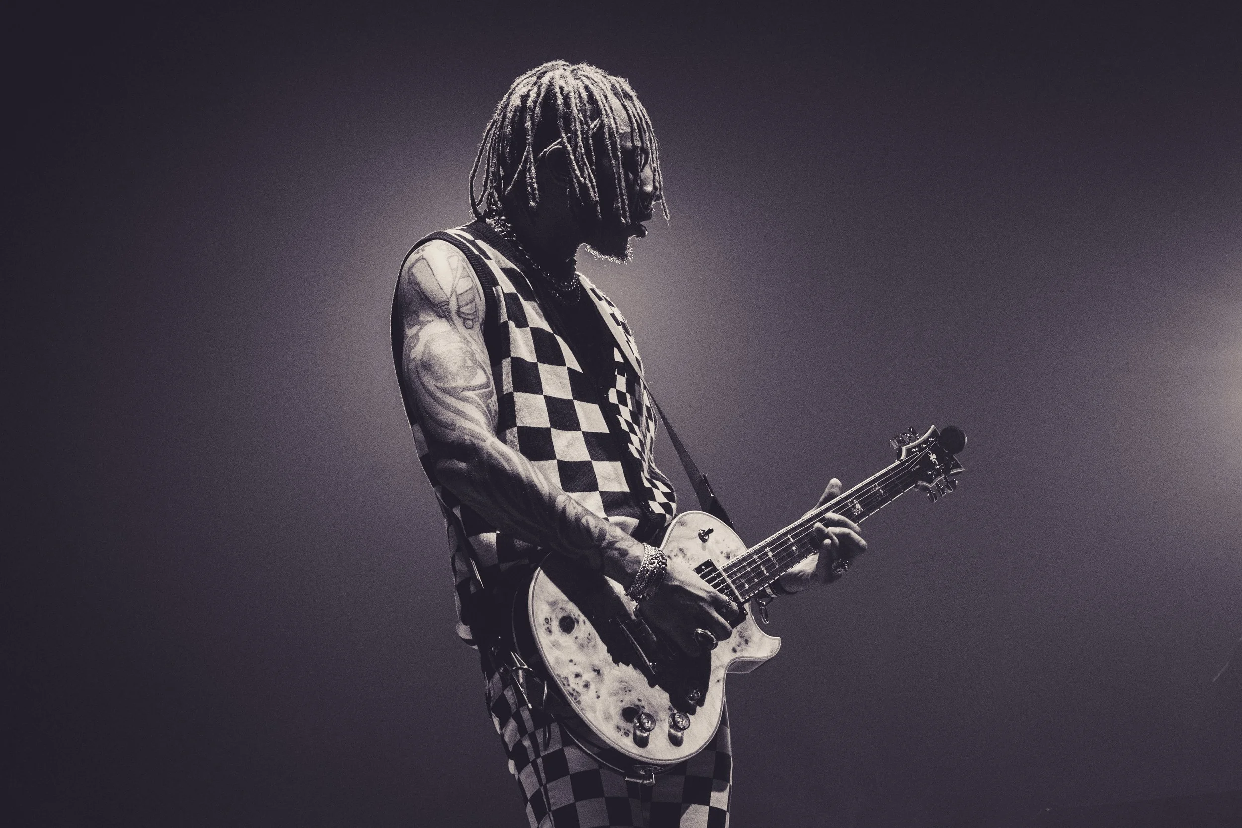 A musician with dreadlocks playing an electric guitar on stage, wearing a sleeveless checkered top and patterned pants, with tattoos on his arms.