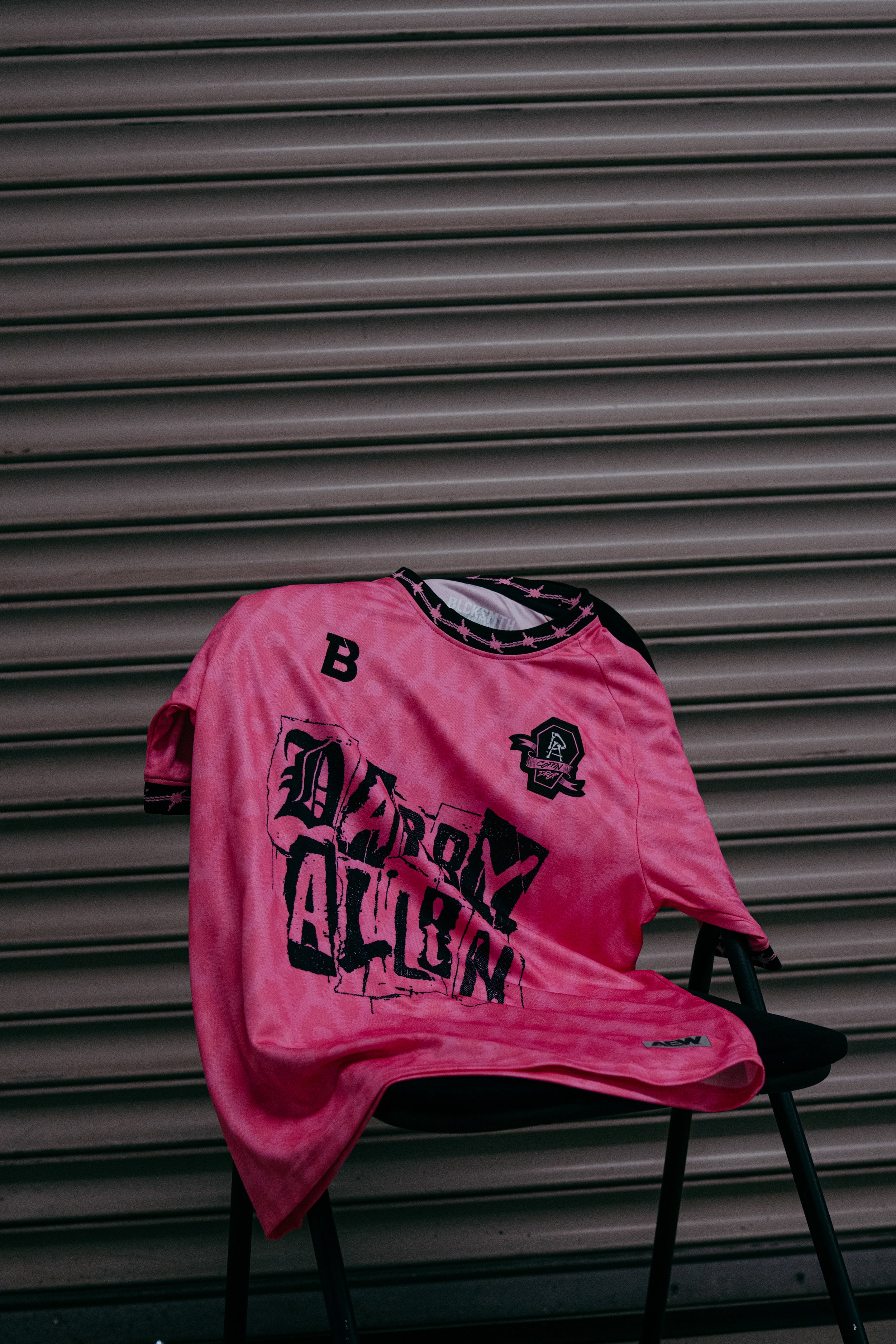 Pink sports jersey with print and patches draped over a black chair in front of a metal roll-up door.