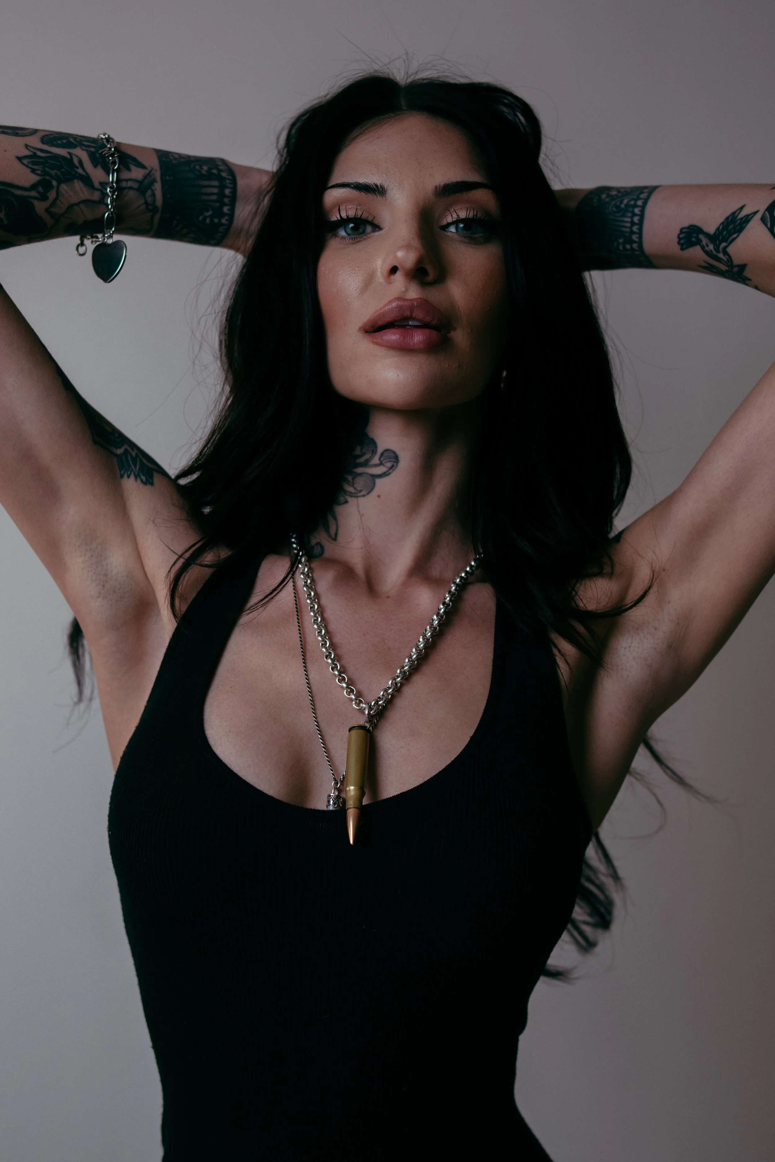 Portrait of a woman with black hair, tattoos on arms, wearing a black tank top and layered necklaces, with arms raised behind her head.