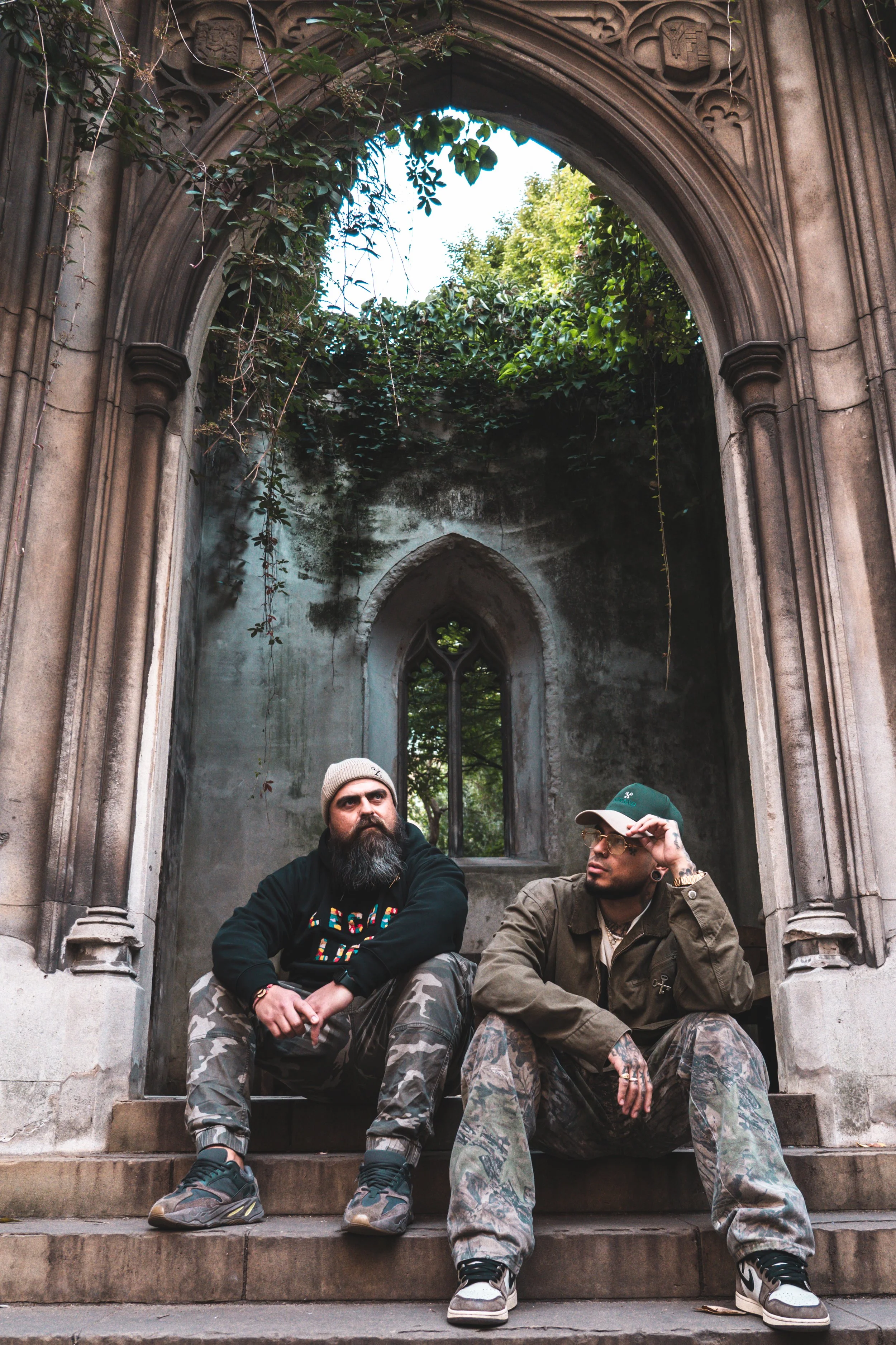 Two men sitting on stone steps in front of an old, Gothic-style stone archway with a small pointed window and overgrown greenery.