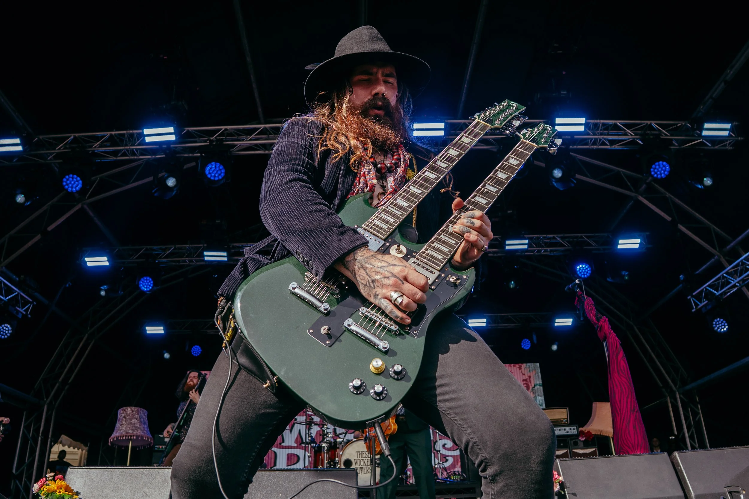 A musician with long hair, beard, dark hat, and necklace playing a double-neck electric guitar on stage during a concert, with stage lighting and band members in the background.