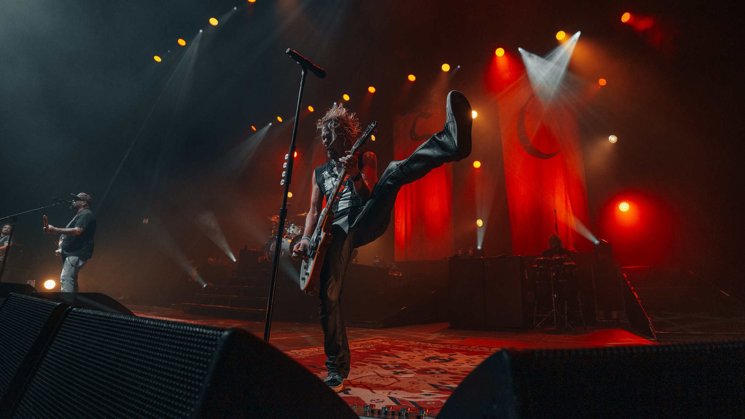 A musician performs on stage with one leg raised high and playing an electric guitar, surrounded by stage lights and other band members.