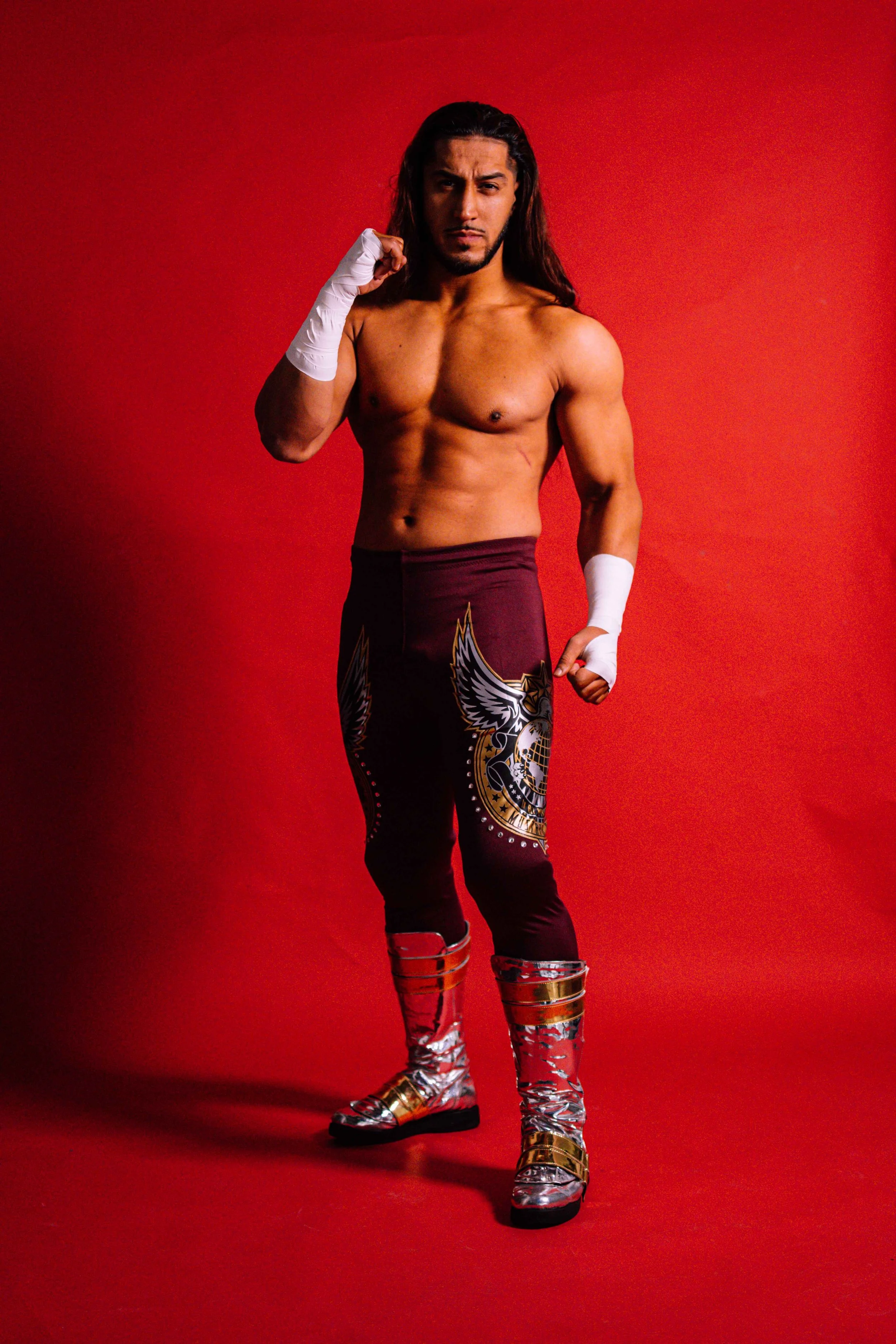 A shirtless man with long dark hair and a serious expression, wearing wrestling gear including colorful tights and metallic boots, posing against a red background.