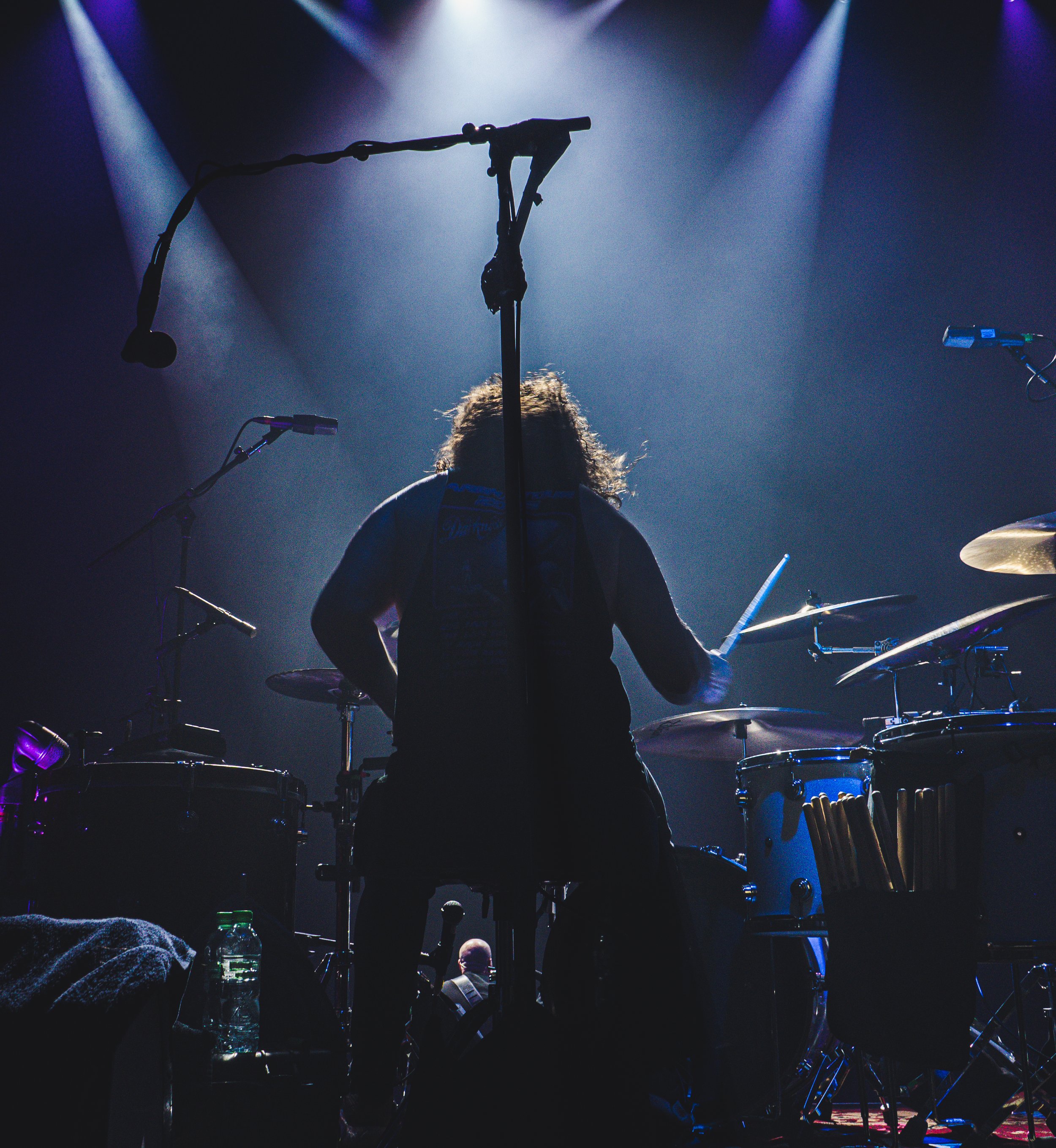 Musician with long curly hair playing drums on stage under purple and blue lighting.