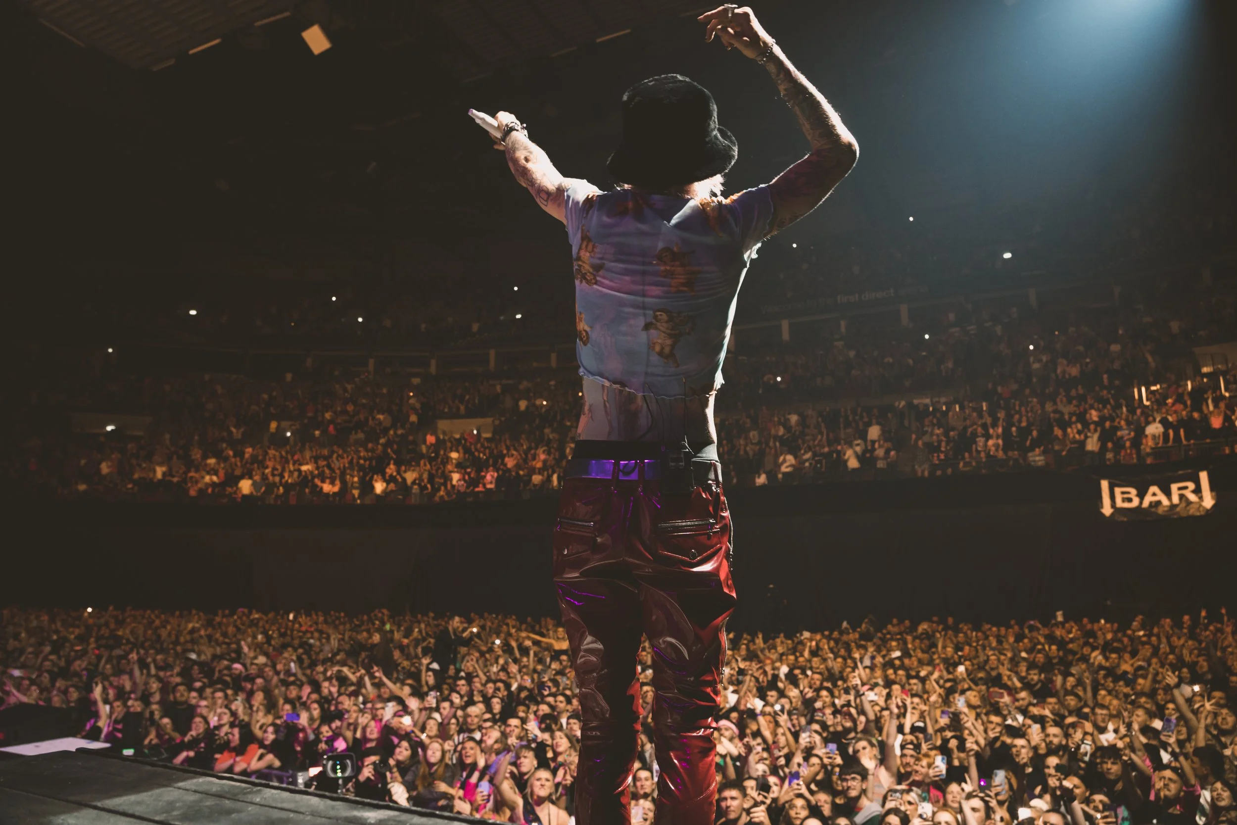Performer on stage facing large audience in concert venue, arms raised, with crowd cheering and taking photos.