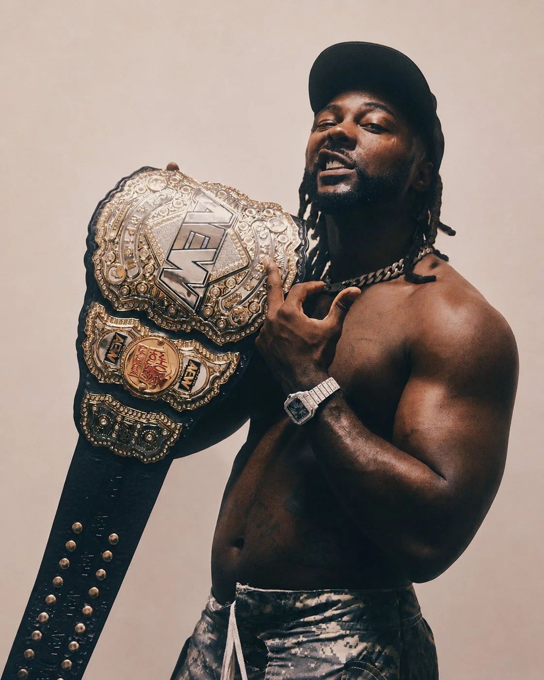 A shirtless man with braided hair wearing a black cap, a chain necklace, and a watch, holding a large championship belt over his shoulder, smiling at the camera.