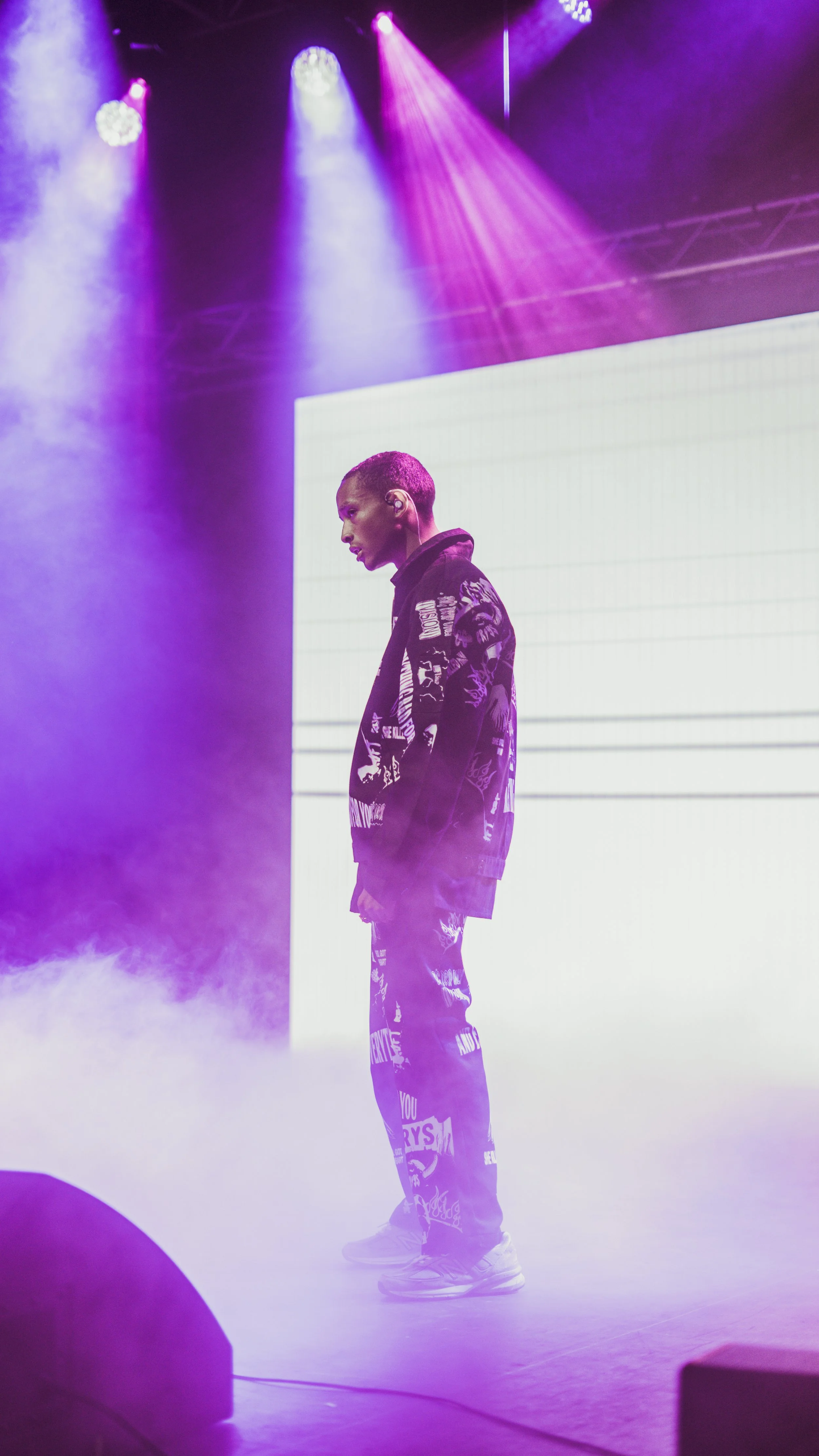 A person standing on stage with purple and pink stage lights and fog, wearing a black jacket and patterned pants.
