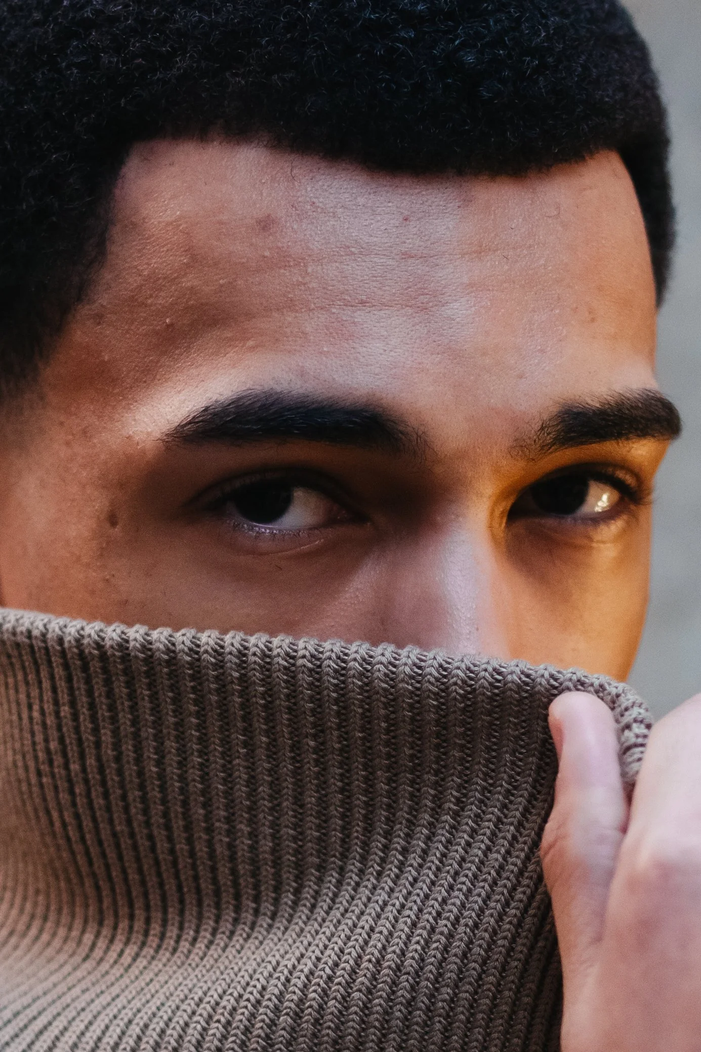 Close-up of a man's face partially hidden behind a brown knit sweater, revealing only his eyes and forehead.