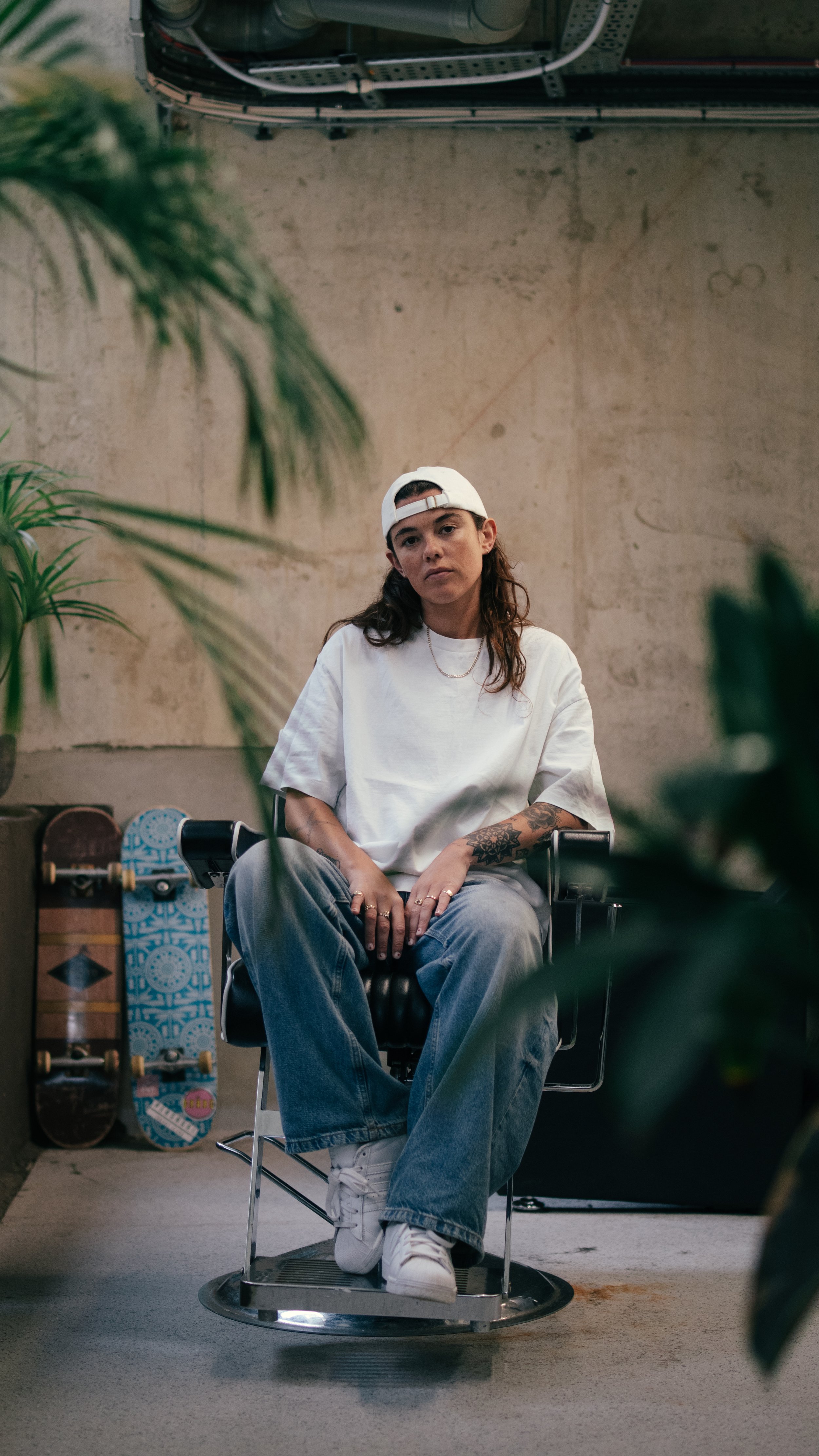 A young woman sitting in a barber chair in a modern space with skateboard decks and plants around her, wearing a white t-shirt, jeans, and a backwards white cap.