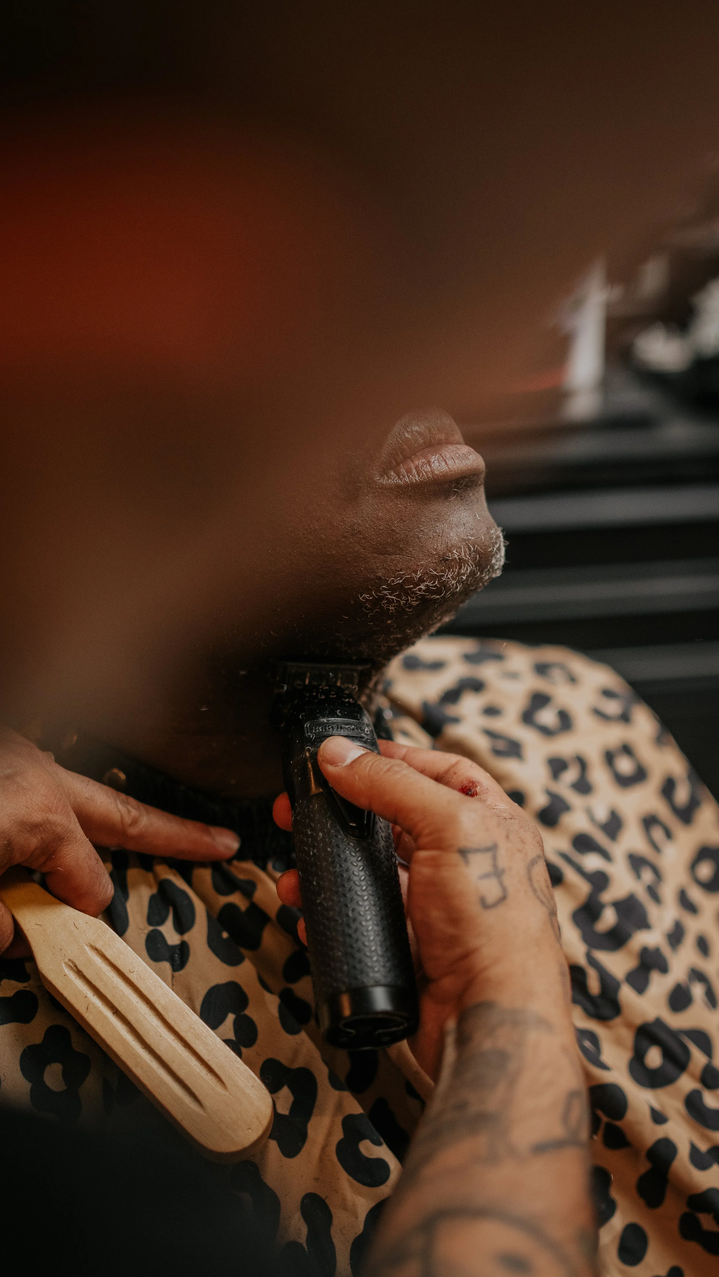 Close-up of person with dark skin getting their beard shaved with electric razor, wearing a leopard print barber cape, with blurred foreground.