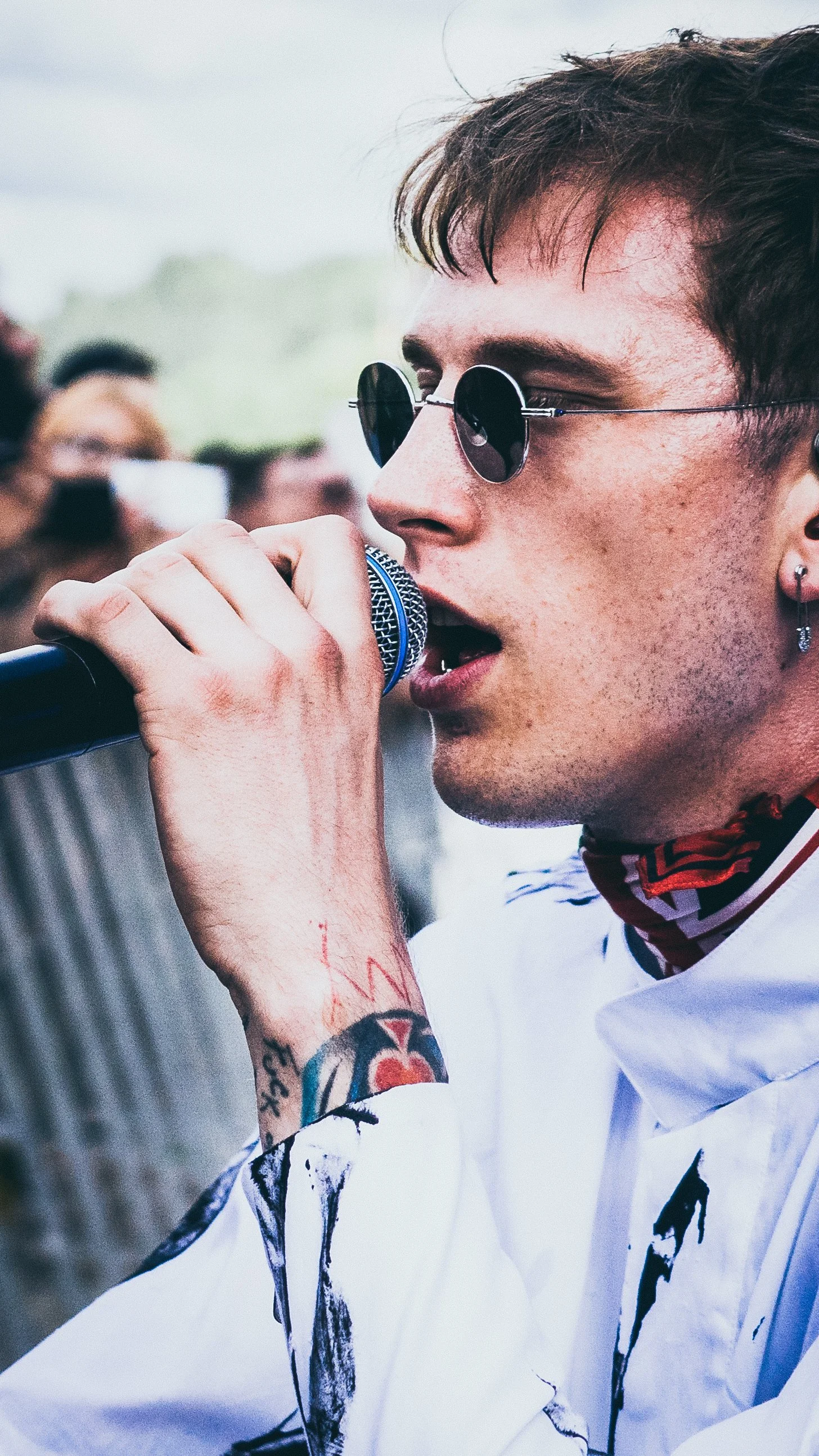 A male singer wearing round sunglasses and earrings, singing into a microphone at an outdoor event, with tattoos on his arm and a crowd in the background.