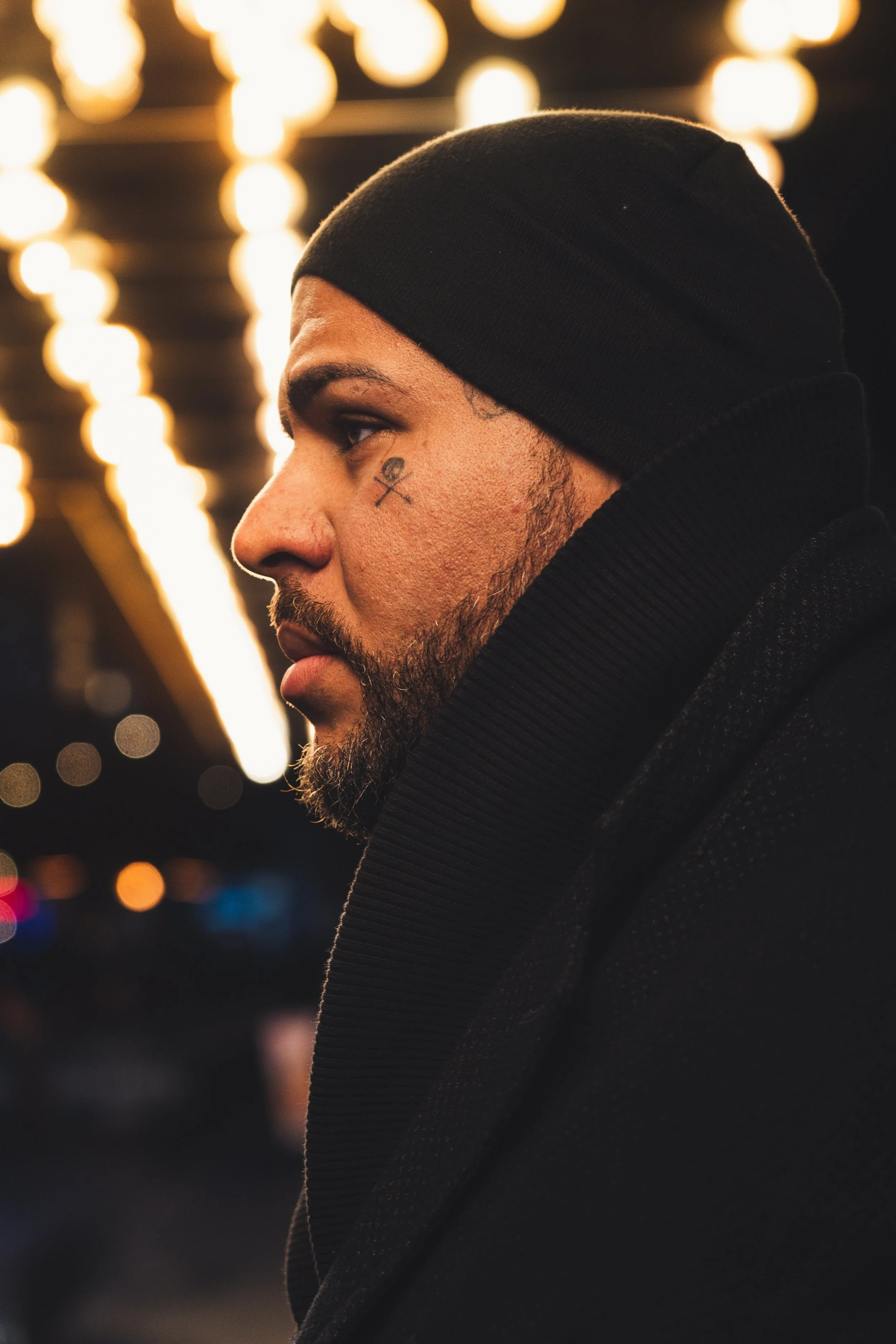 Close-up profile of a man wearing a black beanie and black hoodie, illuminated by warm festival lights in the background.
