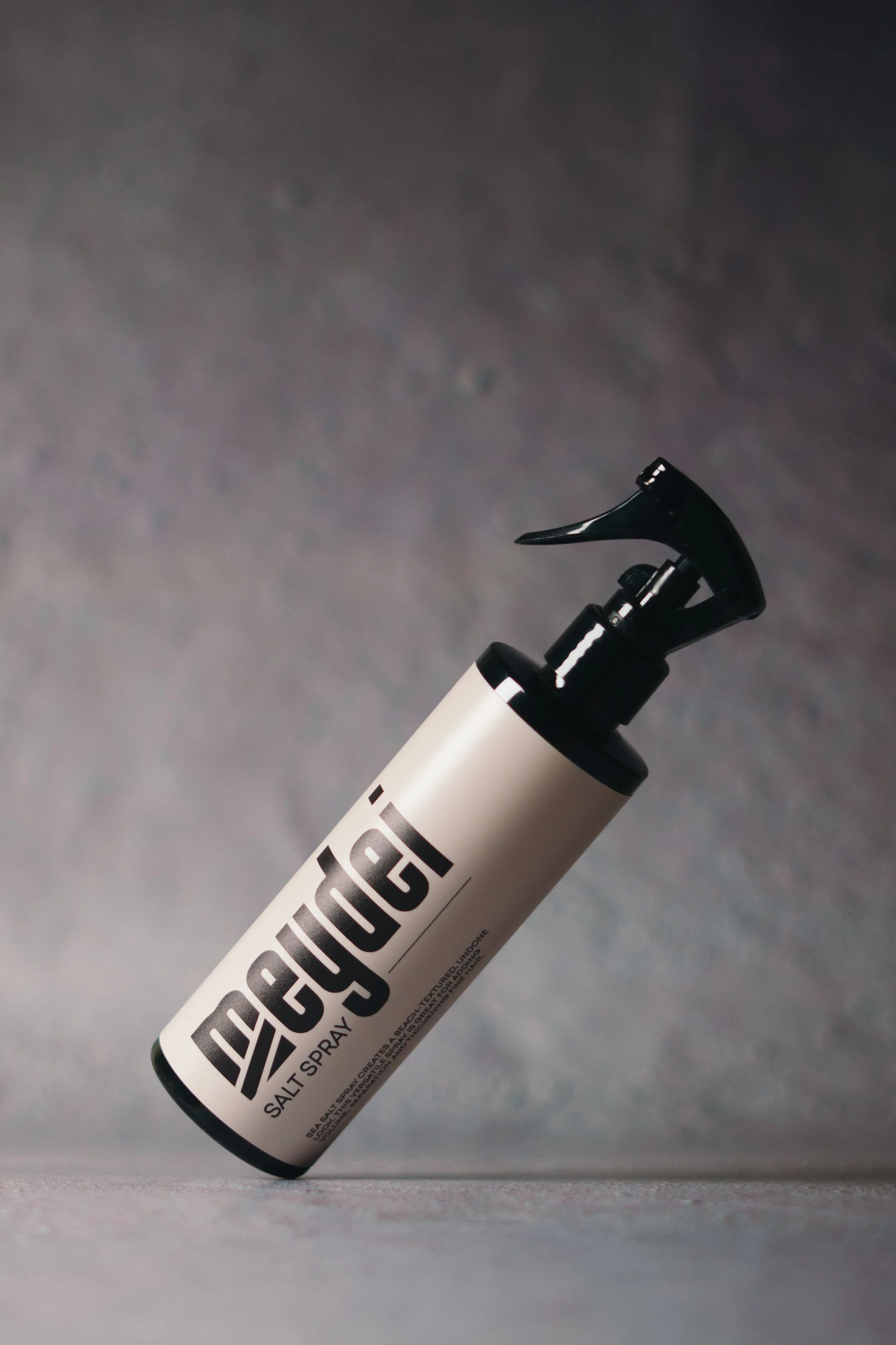 A white bottle of Dehydr Salt Spray with a black pump spray nozzle, positioned at an angle on a gray surface with a blurred gray background.