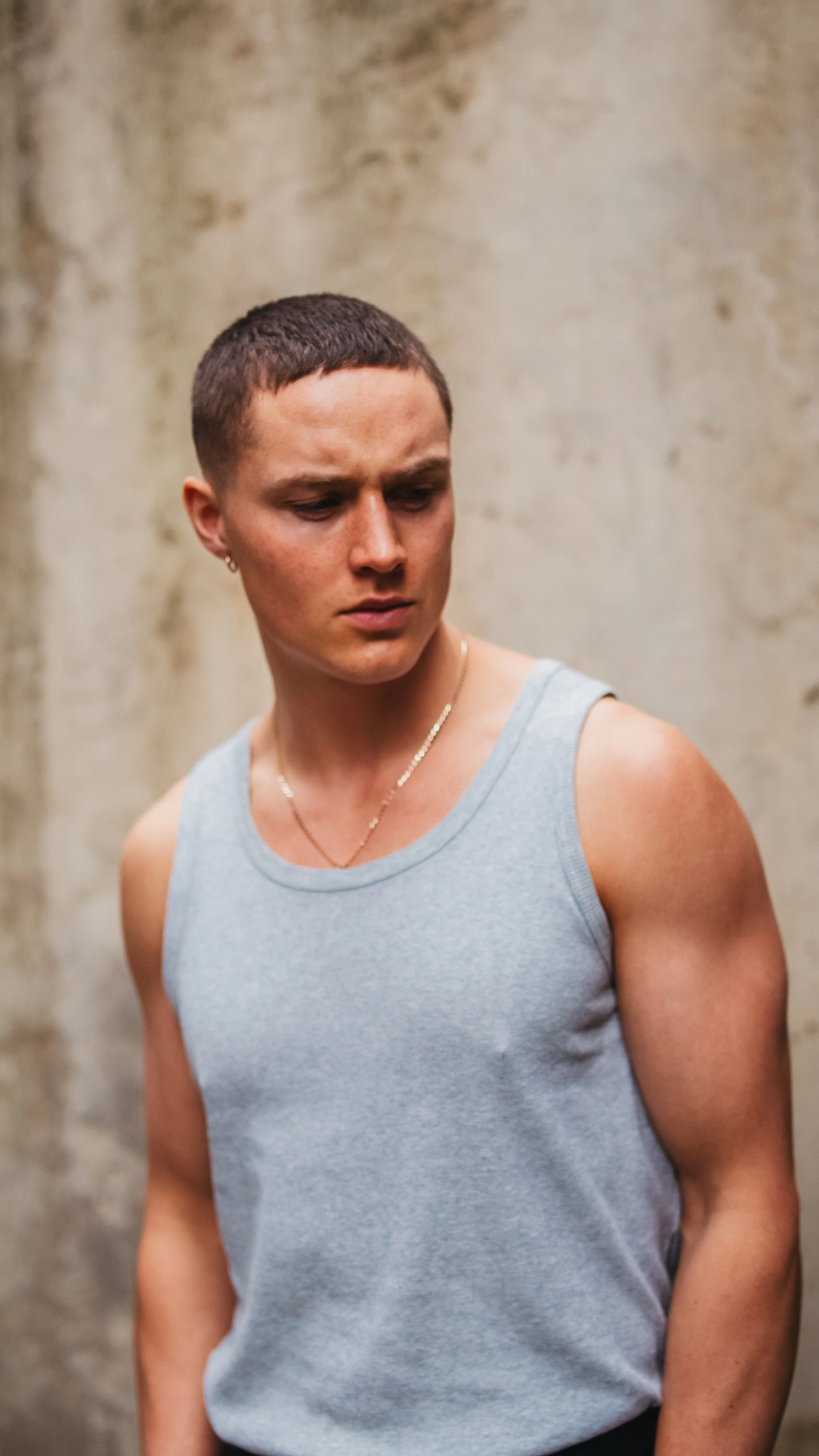 A young person with short hair wearing a sleeveless gray shirt and a gold chain looking to their side against a textured beige wall.