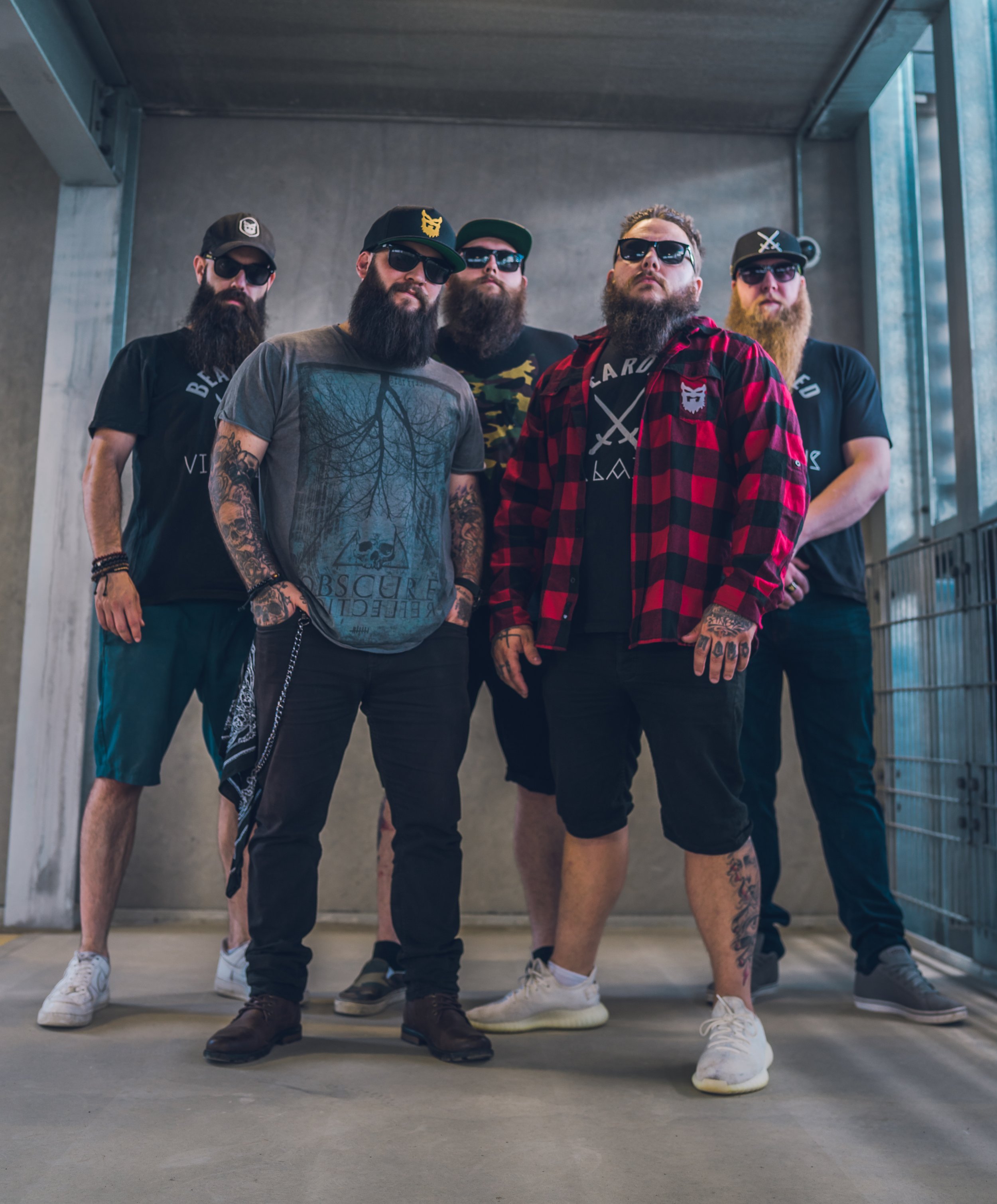 A group of five men with beards, sunglasses, and casual streetwear standing together under an overpass.