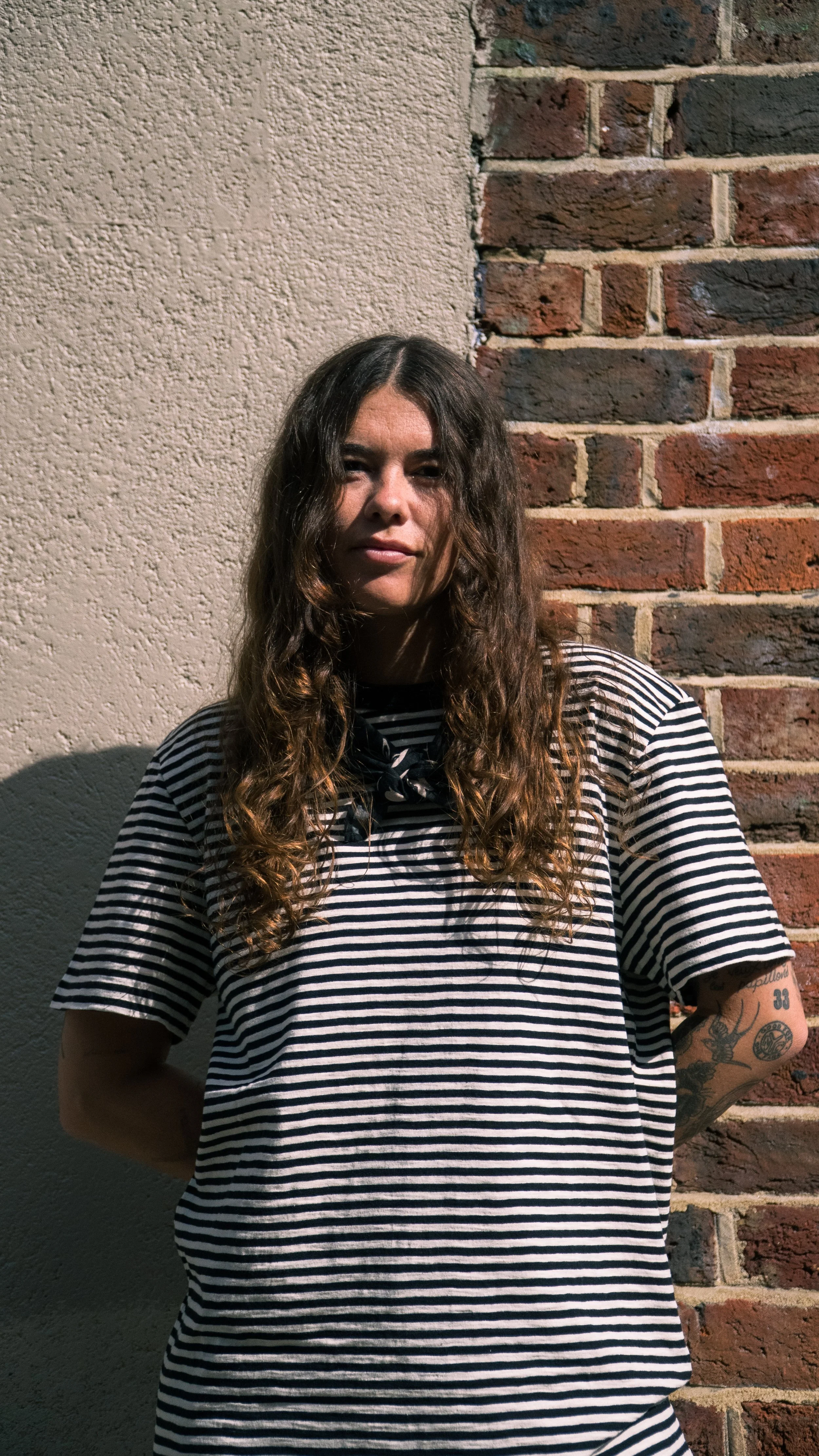 A young woman with long, curly brown hair standing against a wall that is half beige stucco and half red brick, wearing a black and white striped t-shirt and a black neck scarf.