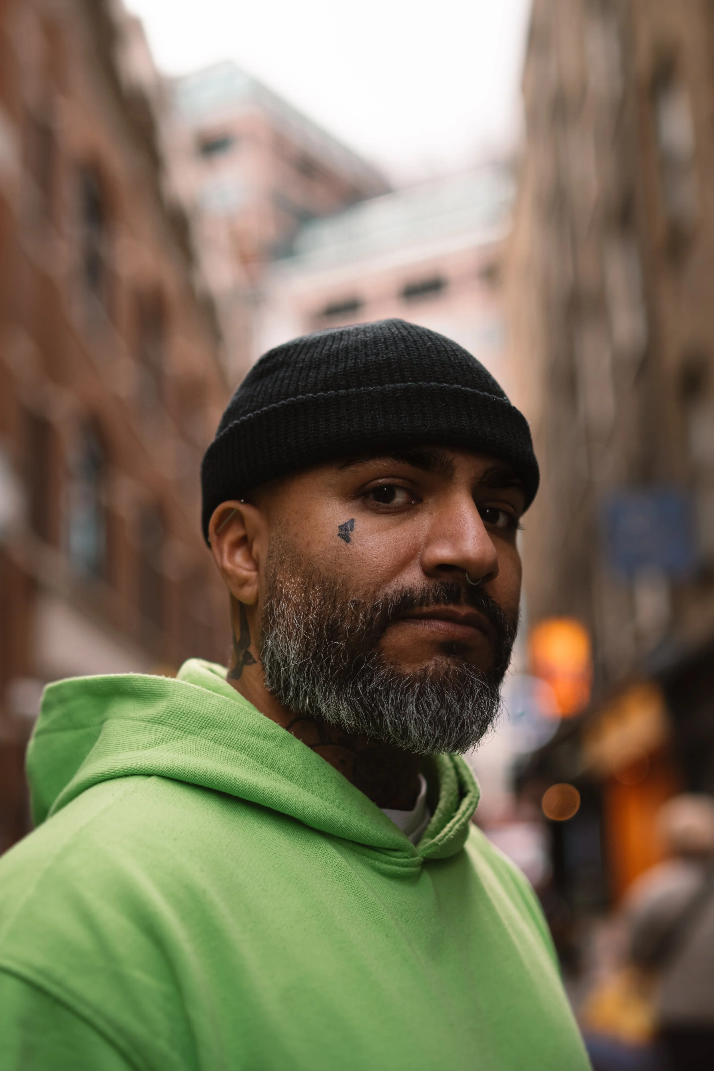 A man with a gray beard and a small tattoo near his eye, wearing a black beanie and a bright green hoodie, stands on a city street with buildings blurred in the background.