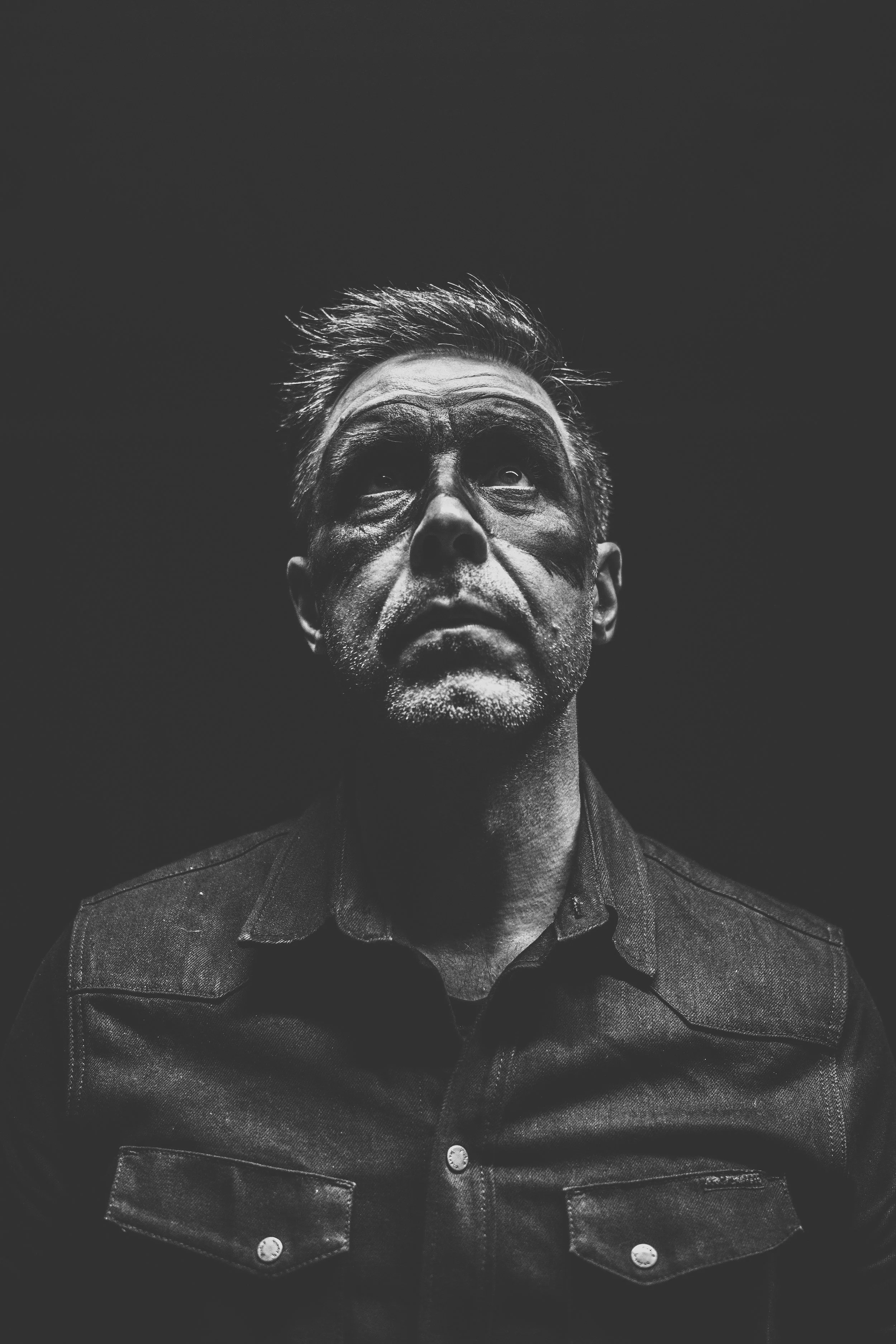 Black and white photo of a man with face paint looking upward, wearing a denim shirt, with a dark background.