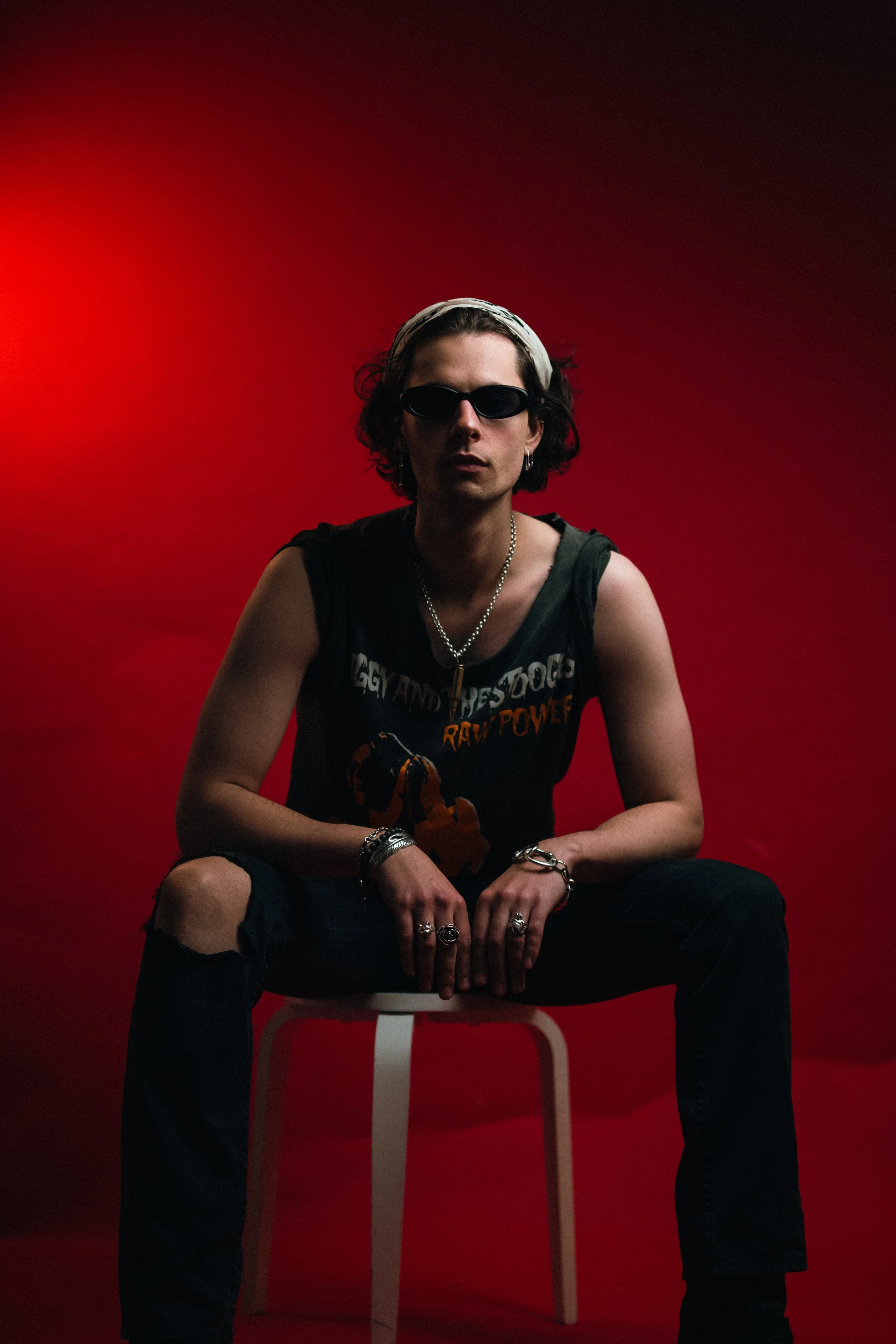 A person with curly dark hair, wearing sunglasses, a sleeveless black T-shirt with graphic text, ripped black jeans, and various jewelry, sitting on a white chair against a red background.