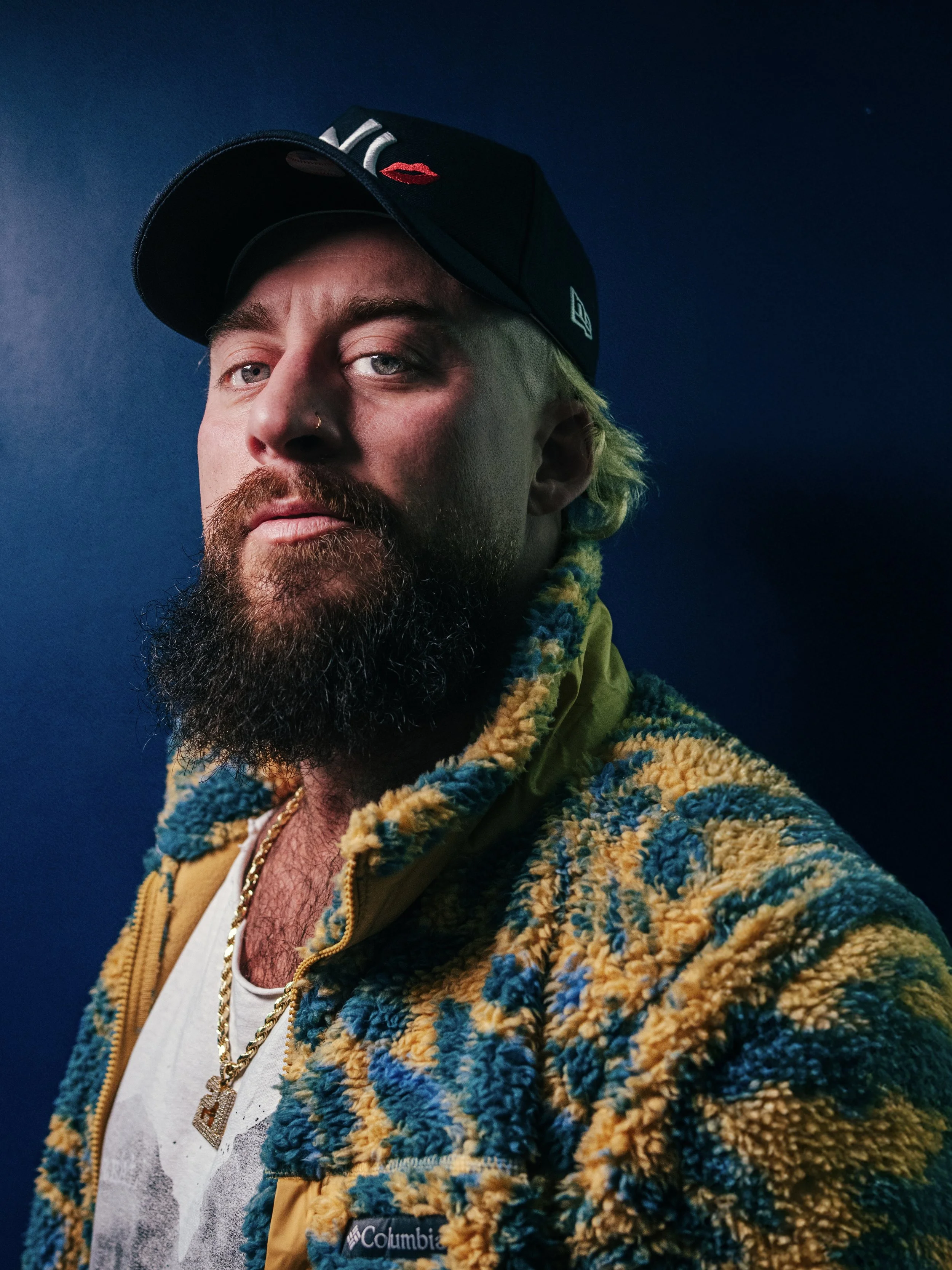 Portrait of a bearded man wearing a black cap with embroidered lips, a patterned fleece jacket, a white t-shirt, and a gold chain with a pendant, against a dark background.