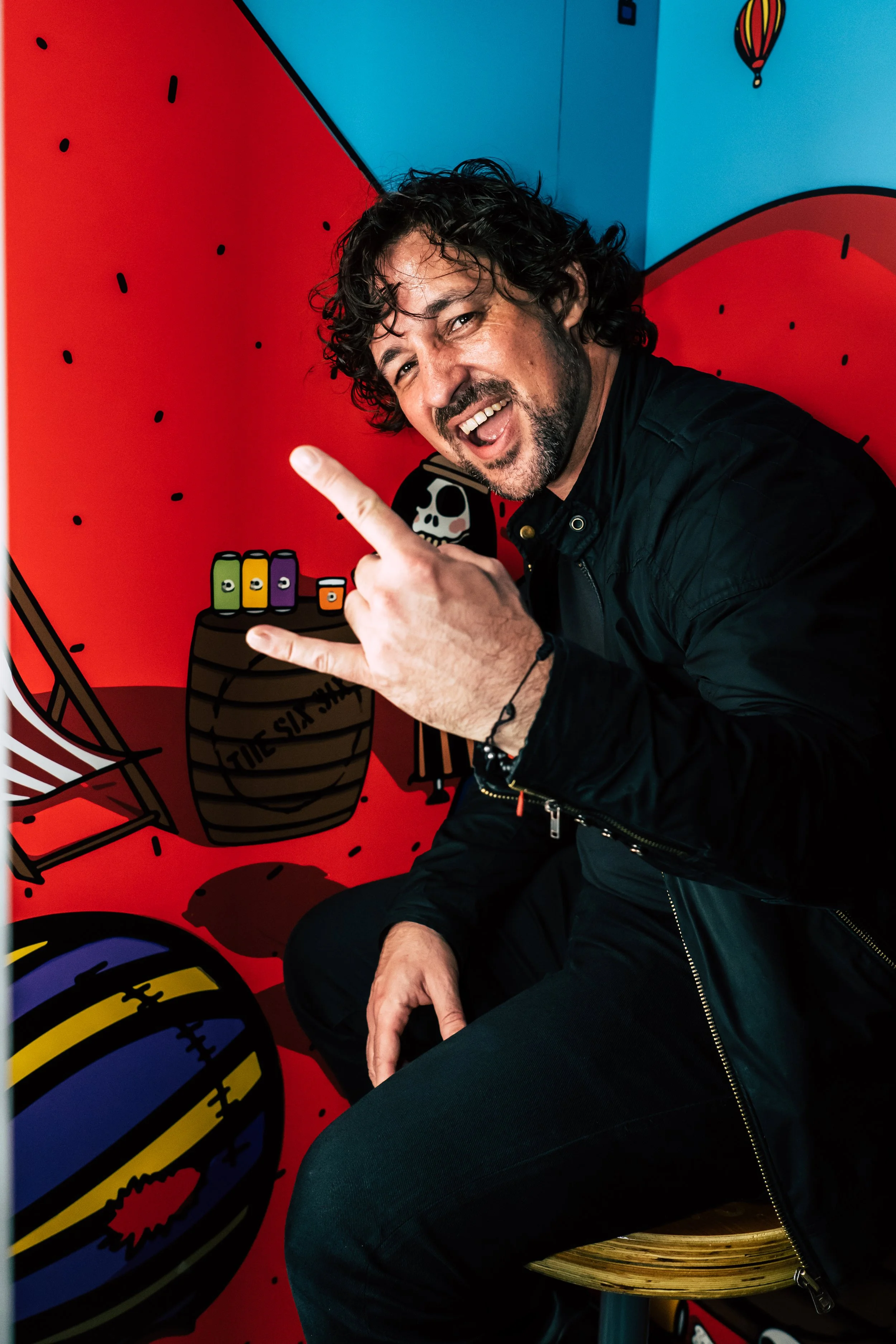A man with dark curly hair and a beard is smiling and making a hand gesture with his right hand, wearing a black jacket, sitting on a wooden stool in front of a colorful cartoon-style backdrop featuring a pirate, a barrel with colored cans, and a bea