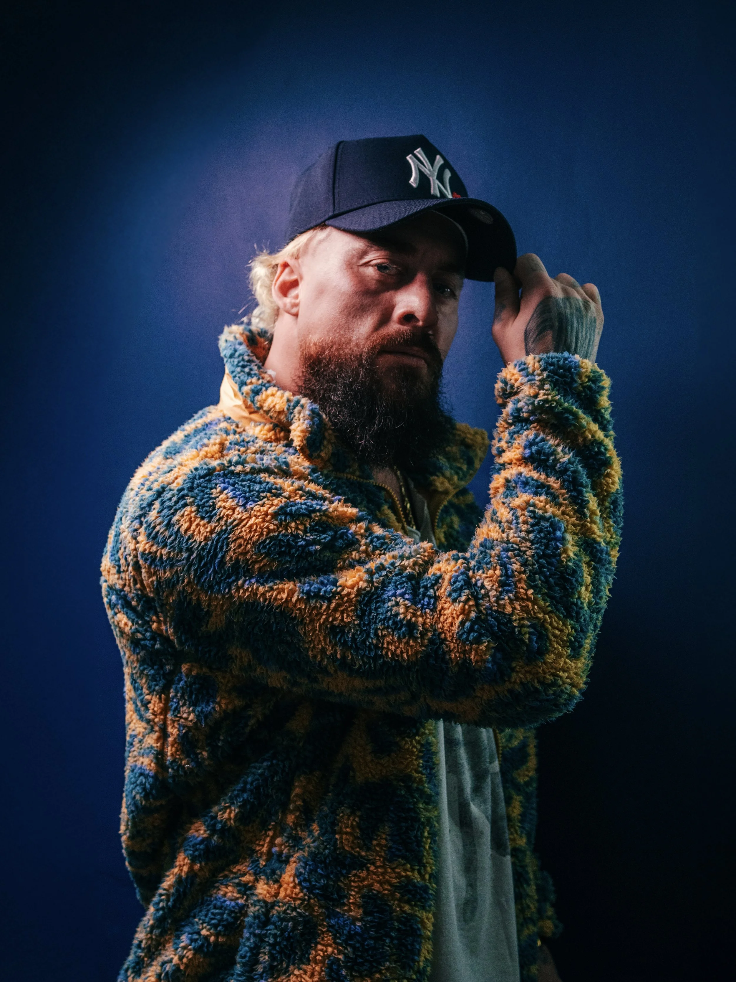 A man with a beard and tattooed hand wearing a New York Yankees cap and a colorful, textured fleece jacket, standing against a dark blue background.