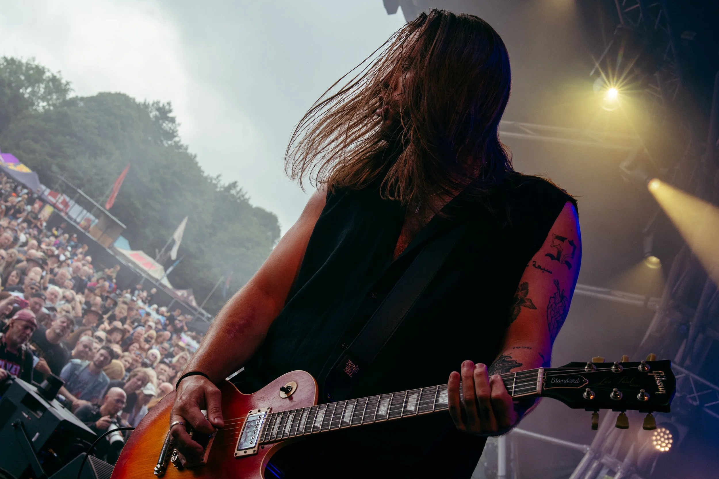 A female guitarist playing electric guitar on stage at a concert, with a large crowd in the background.