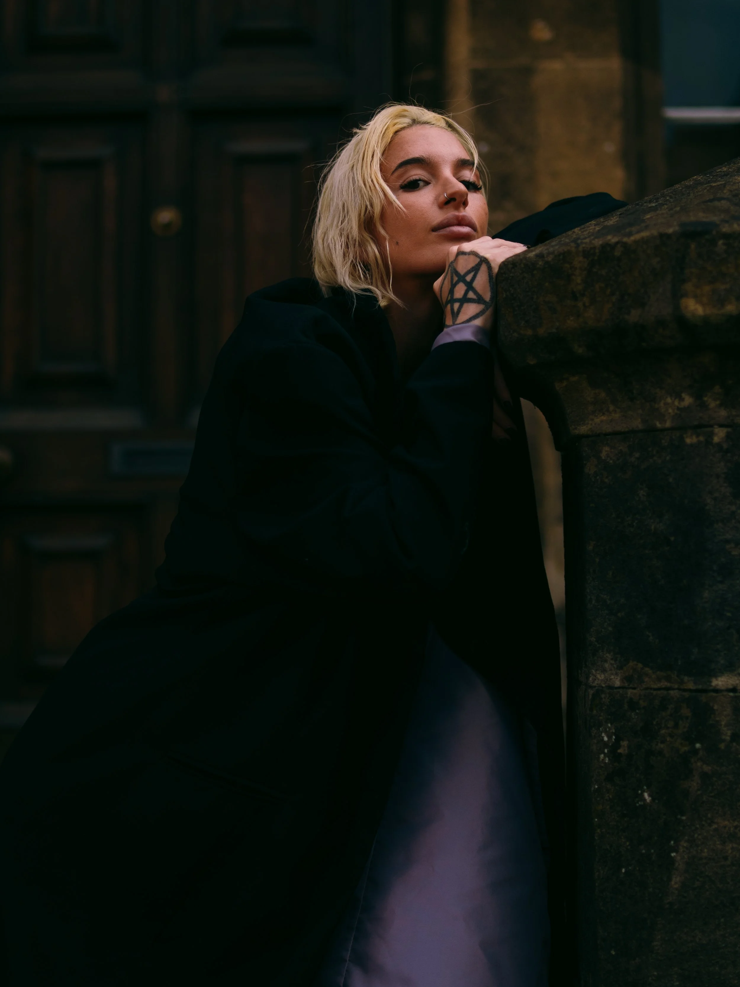 A woman with blonde hair leaning on a stone structure, wearing a black coat. The background shows a dark wooden door and stone wall.