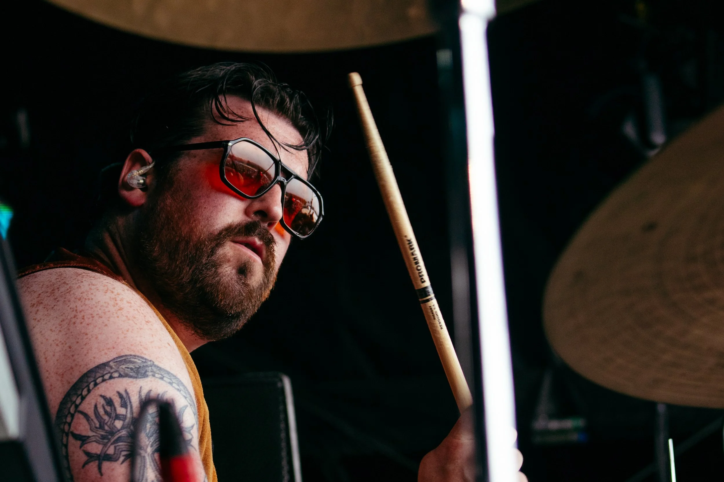 A bearded man wearing sunglasses playing drums, holding a drumstick, with a tattoo on his arm.