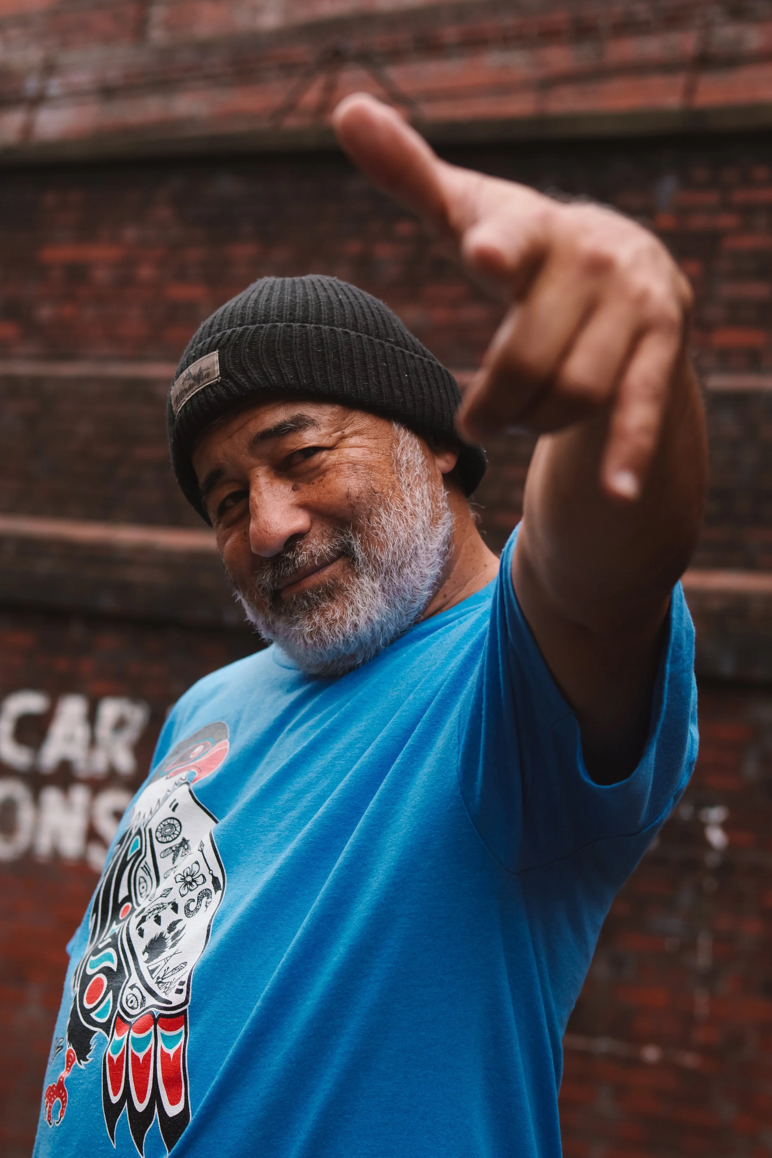 A man with a gray beard wearing a black beanie and a blue t-shirt, pointing towards the camera with a confident expression, standing in front of a brick wall.