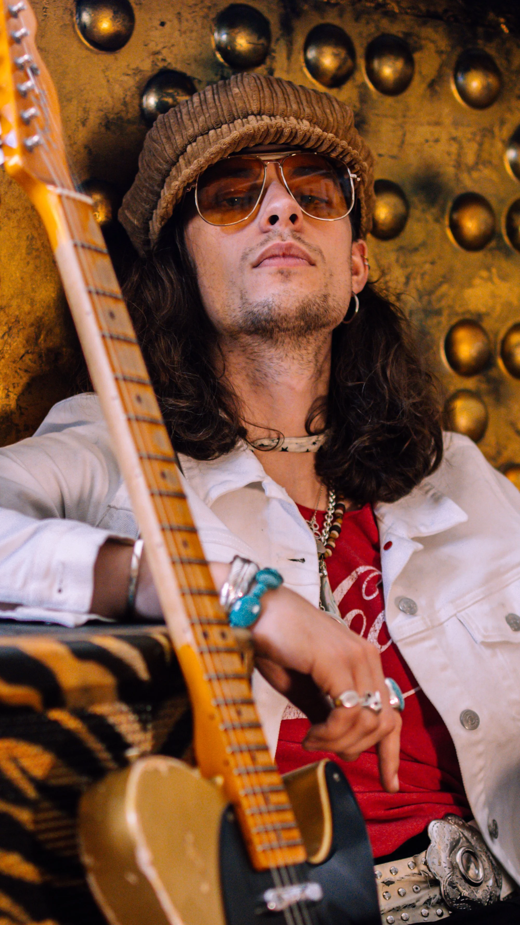 A man with long dark hair, wearing sunglasses, a brown knit hat, and a white jacket, sitting in front of a gold wall with round metallic decorations, holding a guitar.